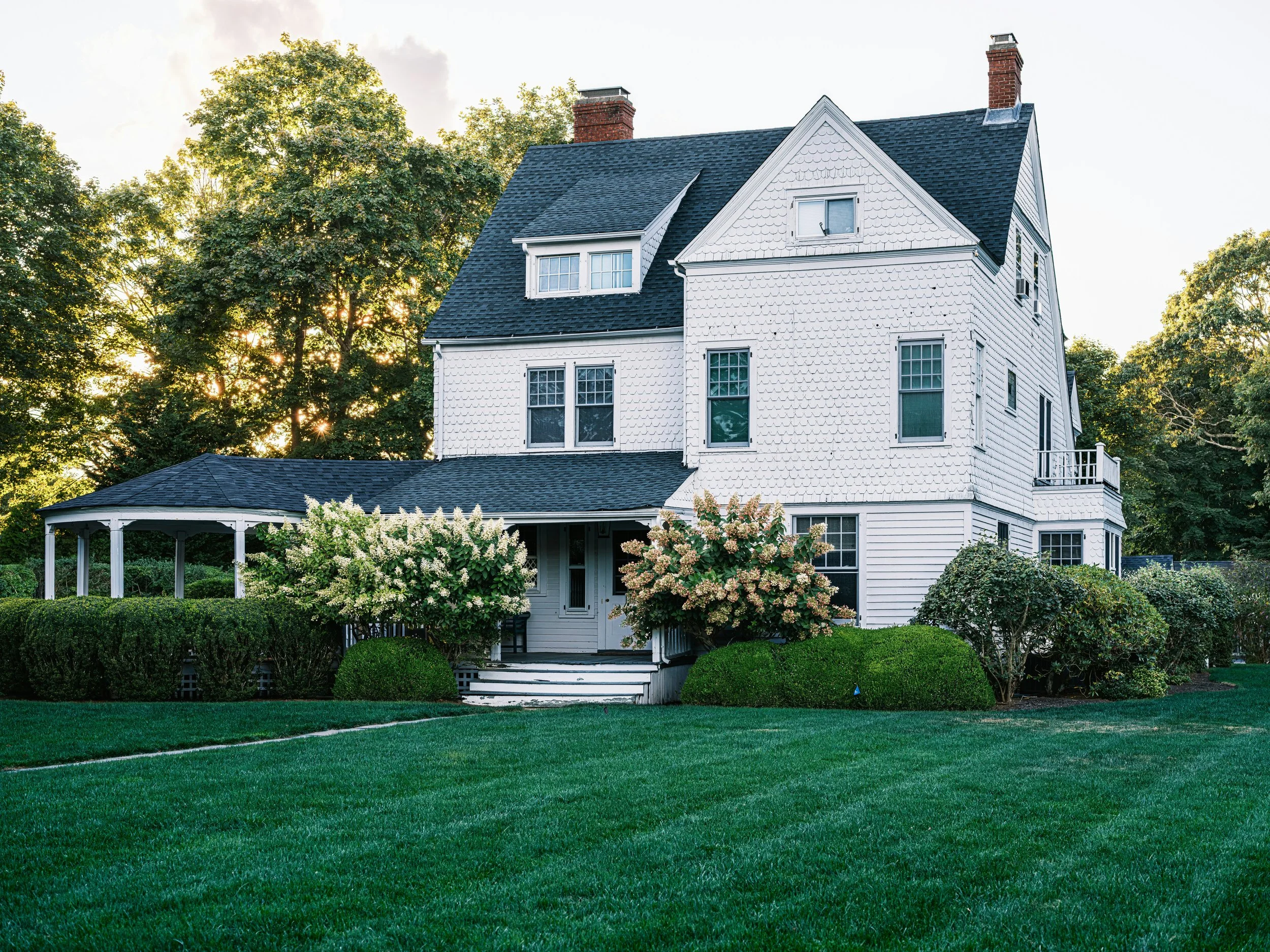 A large white house with a dark shingle roof, multiple windows, and a small balcony on the side. The house has a front porch, well-maintained greenery, bushes, and a lush, green lawn. Tall trees are in the background under a partly cloudy sky.