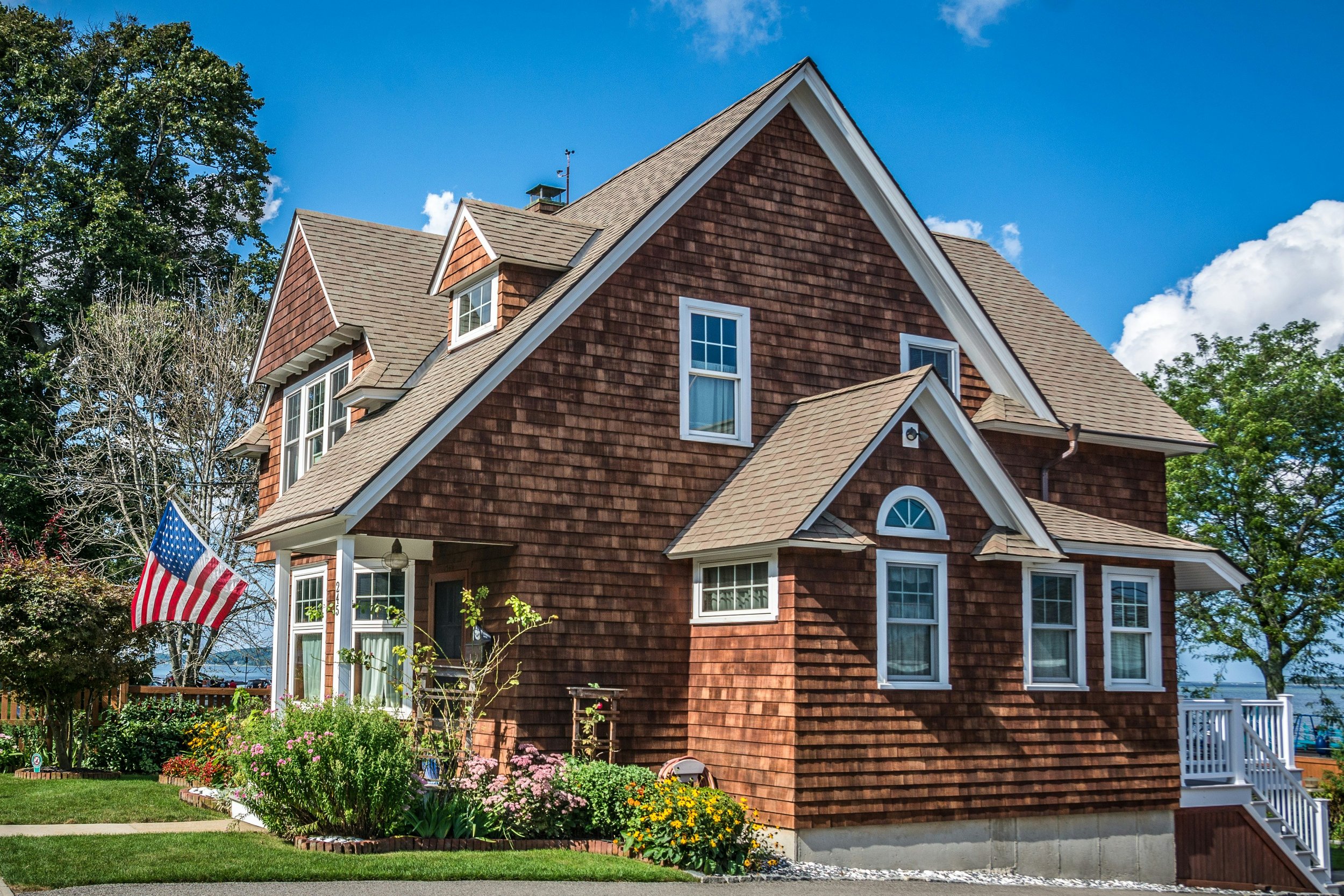 A large house with brown wooden shingles, gabled roof, multiple windows, front porch, and garden with colorful flowers, American flag, and green lawn, under a blue sky with some clouds.