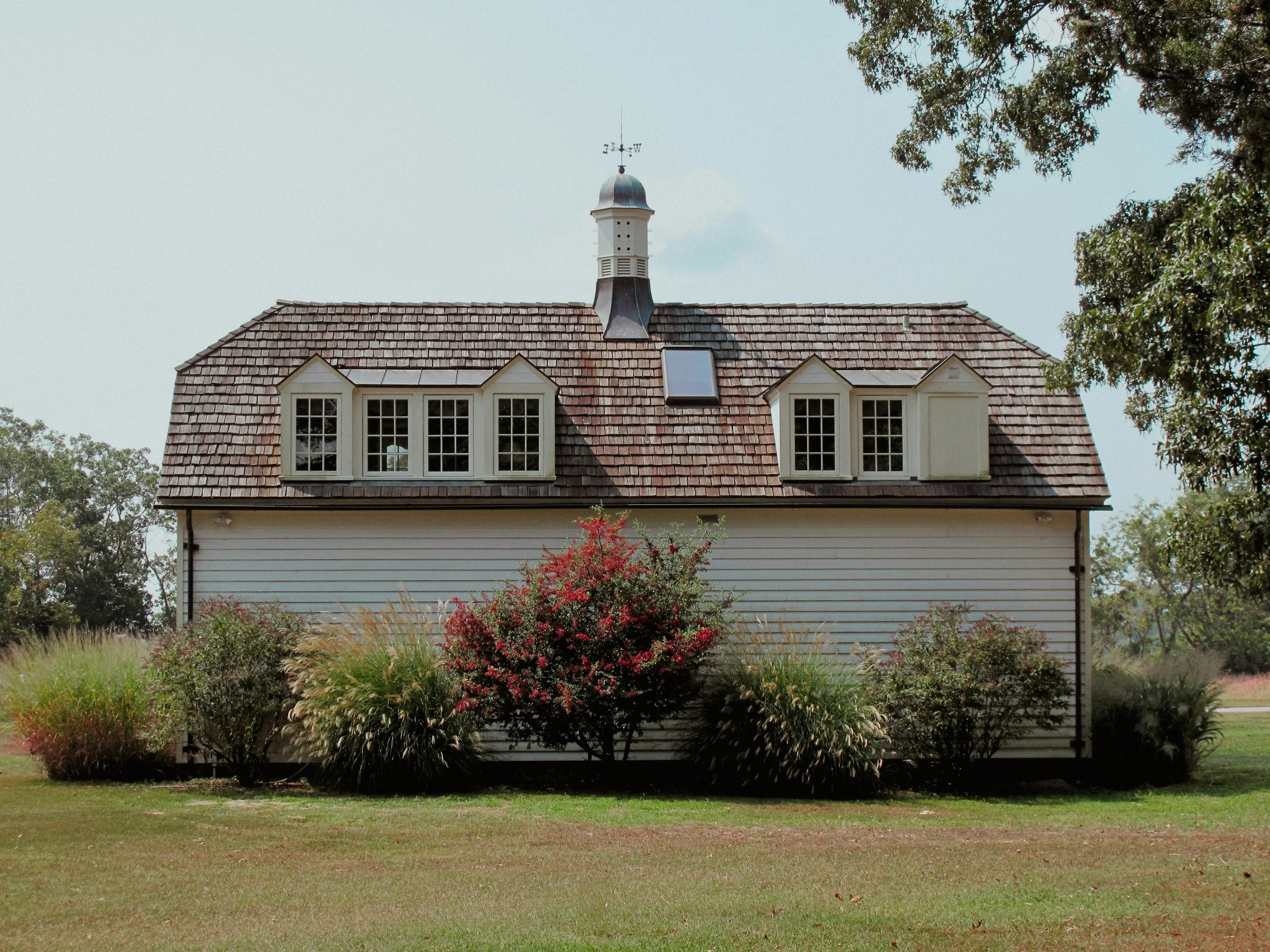 Front view of a house with a steep roof, dormer windows, and a cupola with weather vane on top, surrounded by trees and a well-maintained lawn.