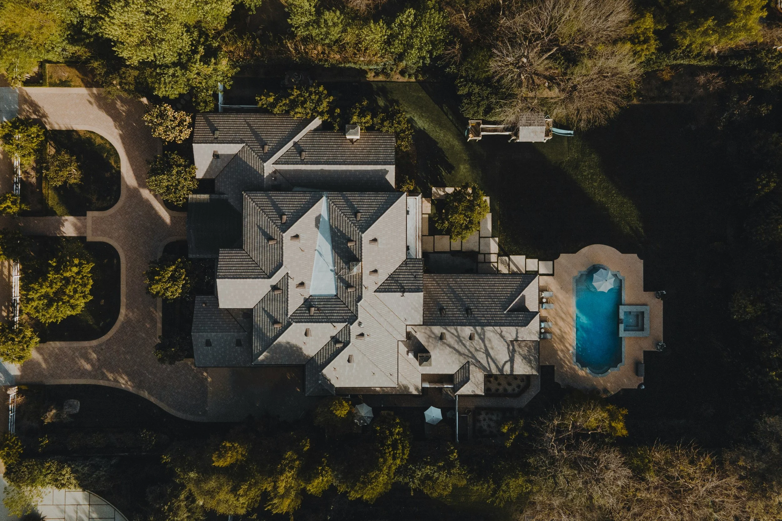 An aerial view of a large house with a gray roof, surrounding trees, a swimming pool, and a brick driveway.