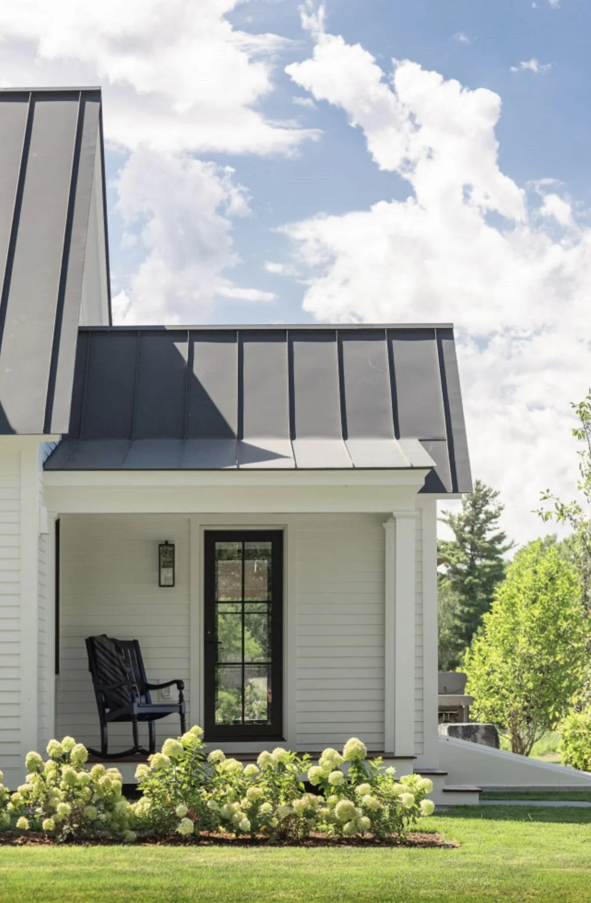 A modern house with a black metal roof, white siding, a porch with black rocking chairs, and a garden with white flowers, trees, and a blue sky with white clouds.