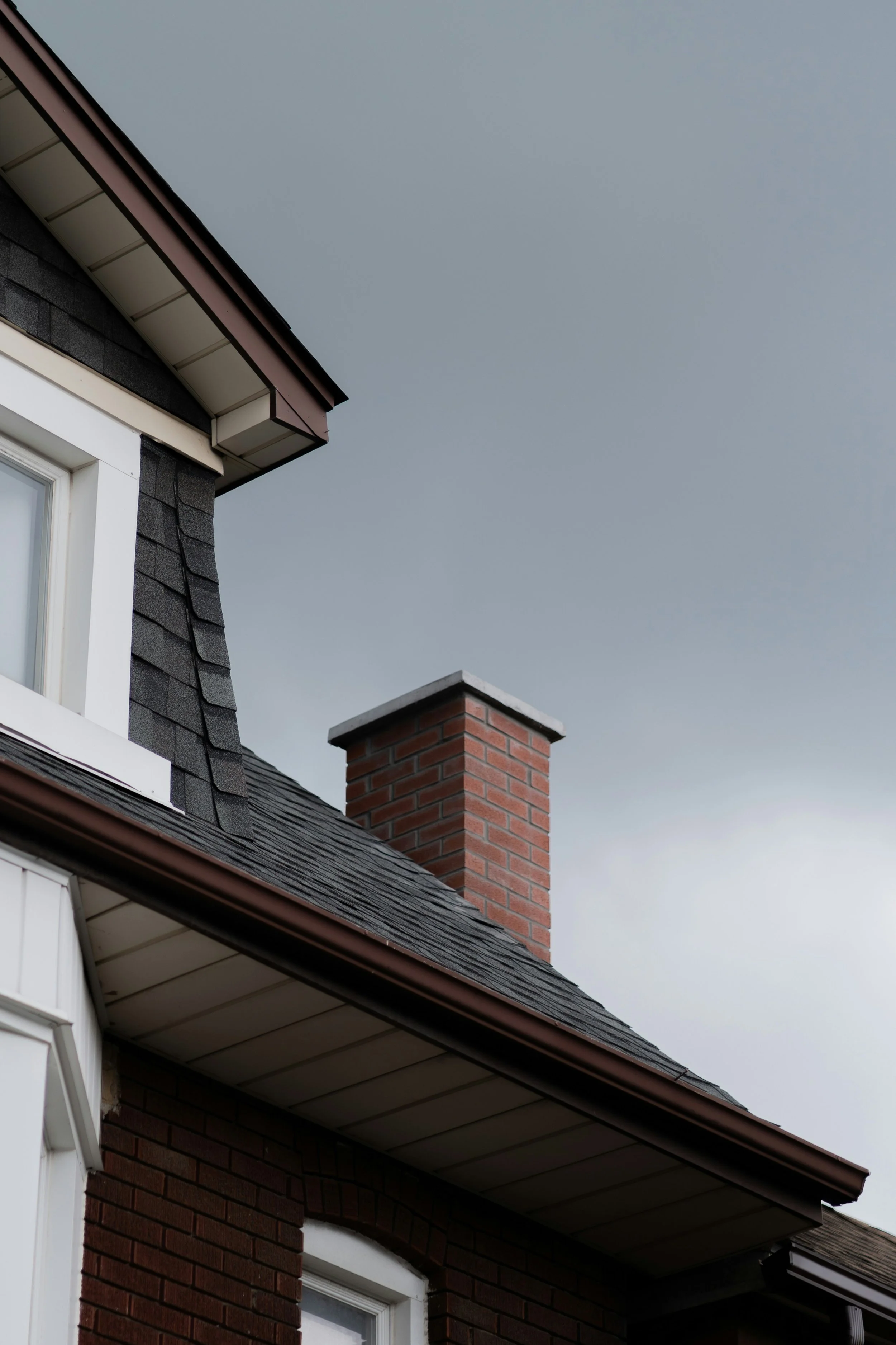 Close-up of the corner of a house with dark shingle siding, a brick chimney, a white-framed window, and an overcast sky in the background.