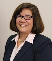 A professional woman with shoulder-length dark hair, glasses, and a warm smile, wearing a navy blazer and white blouse.