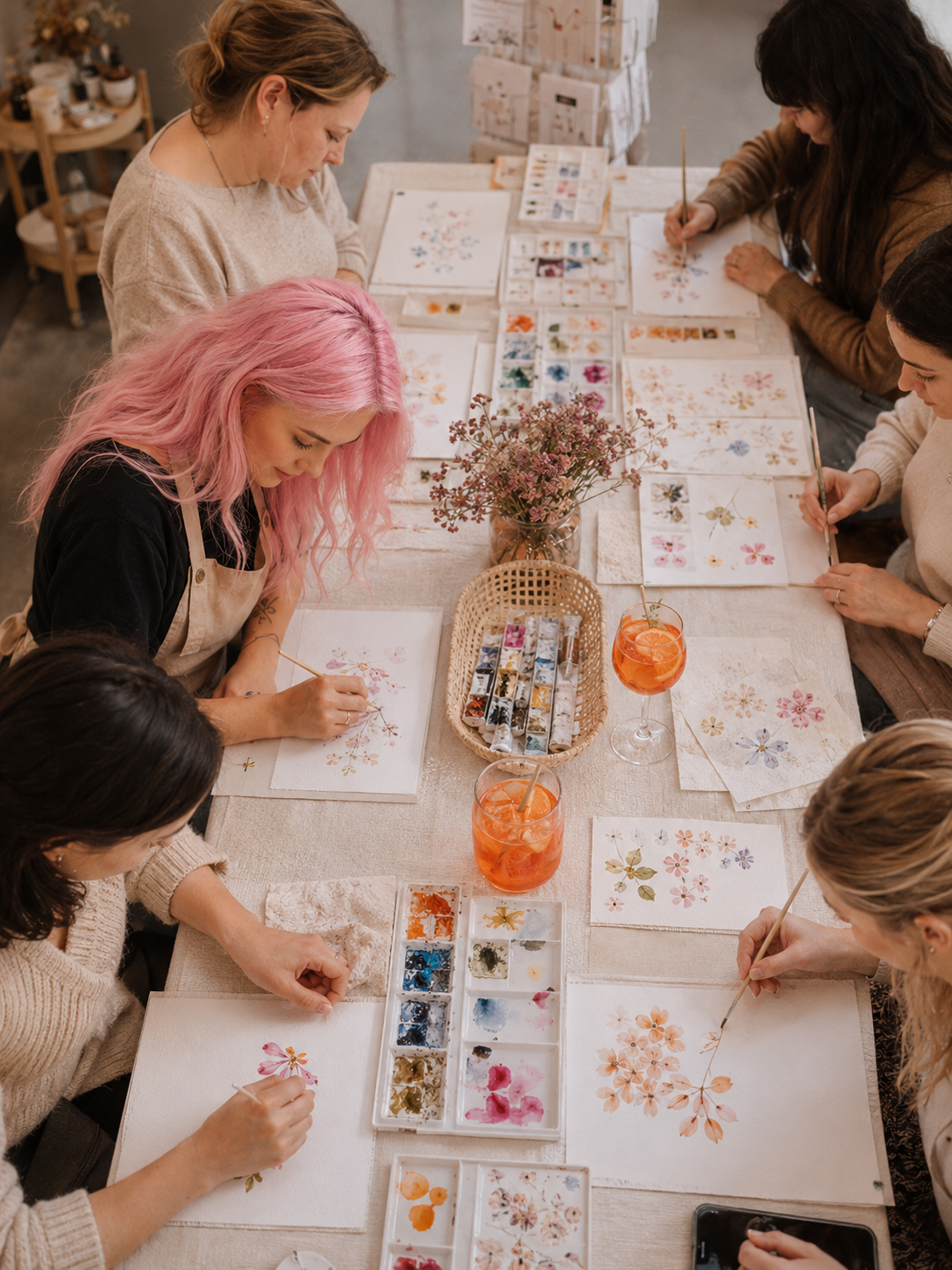 Menschen beim Aquarellmalen mit Blumenmotiven in einem Atelier, zusammen an einem Tisch mit Malmaterial und Getränken.