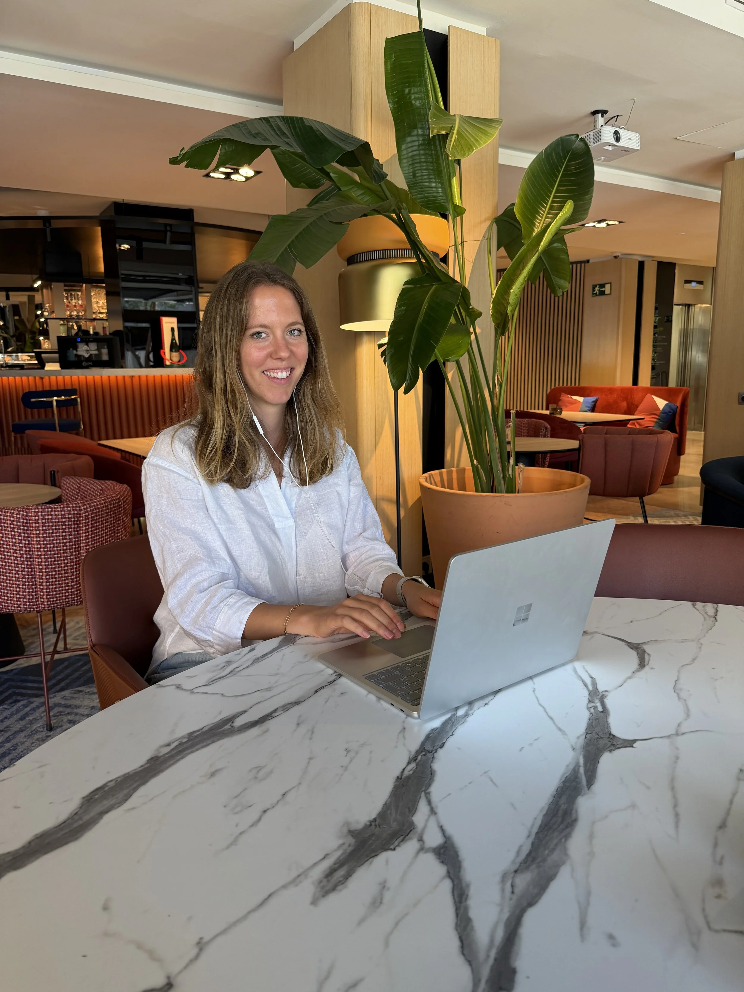A woman with long blonde hair, wearing a white shirt, sitting at a marble table with a silver laptop, in a modern interior with green plants and colorful seating.