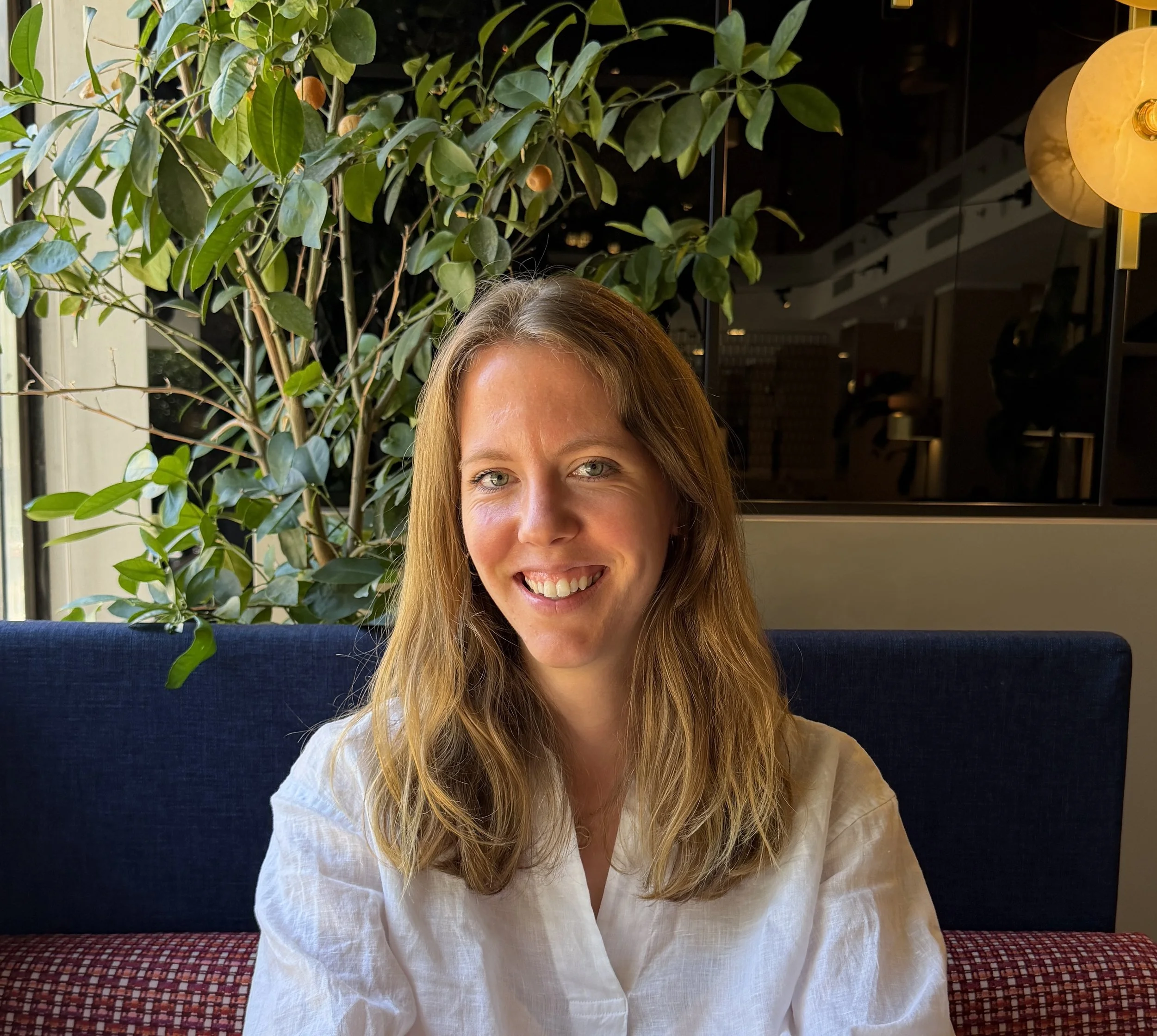 Close-up of a smiling woman with long, light brown hair, wearing a white shirt, sitting at a table indoors with a large leafy plant and warm lighting in the background.