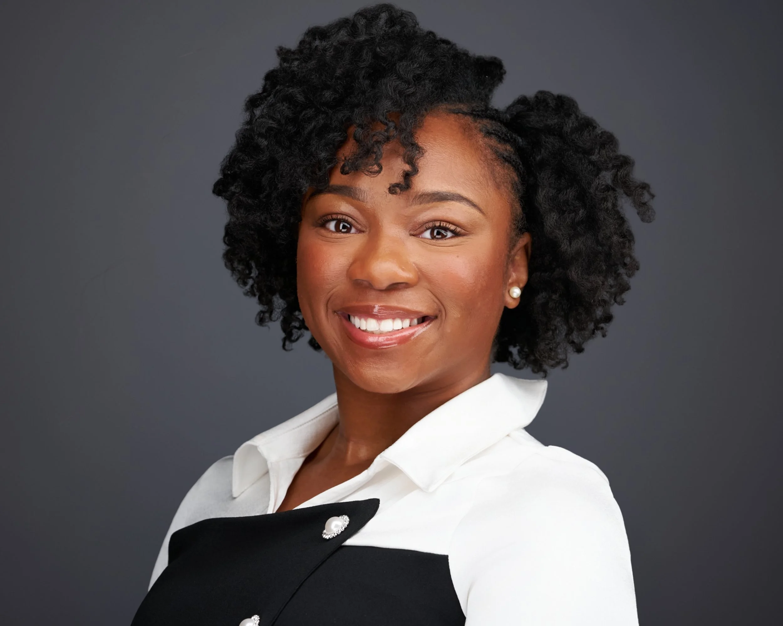 Portrait of an African American woman with natural curly hair, wearing a white shirt, and smiling, set against a dark background.