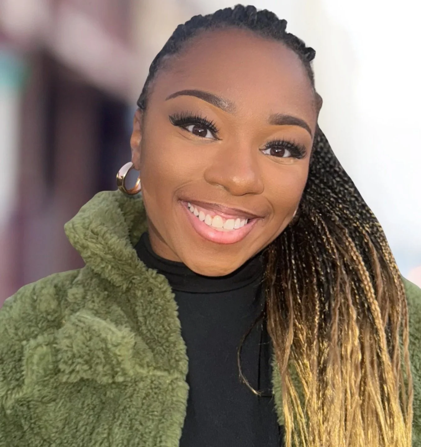 A young woman with braided hair and large earrings smiling warmly, wearing a black top and green fuzzy jacket, with a blurred background.