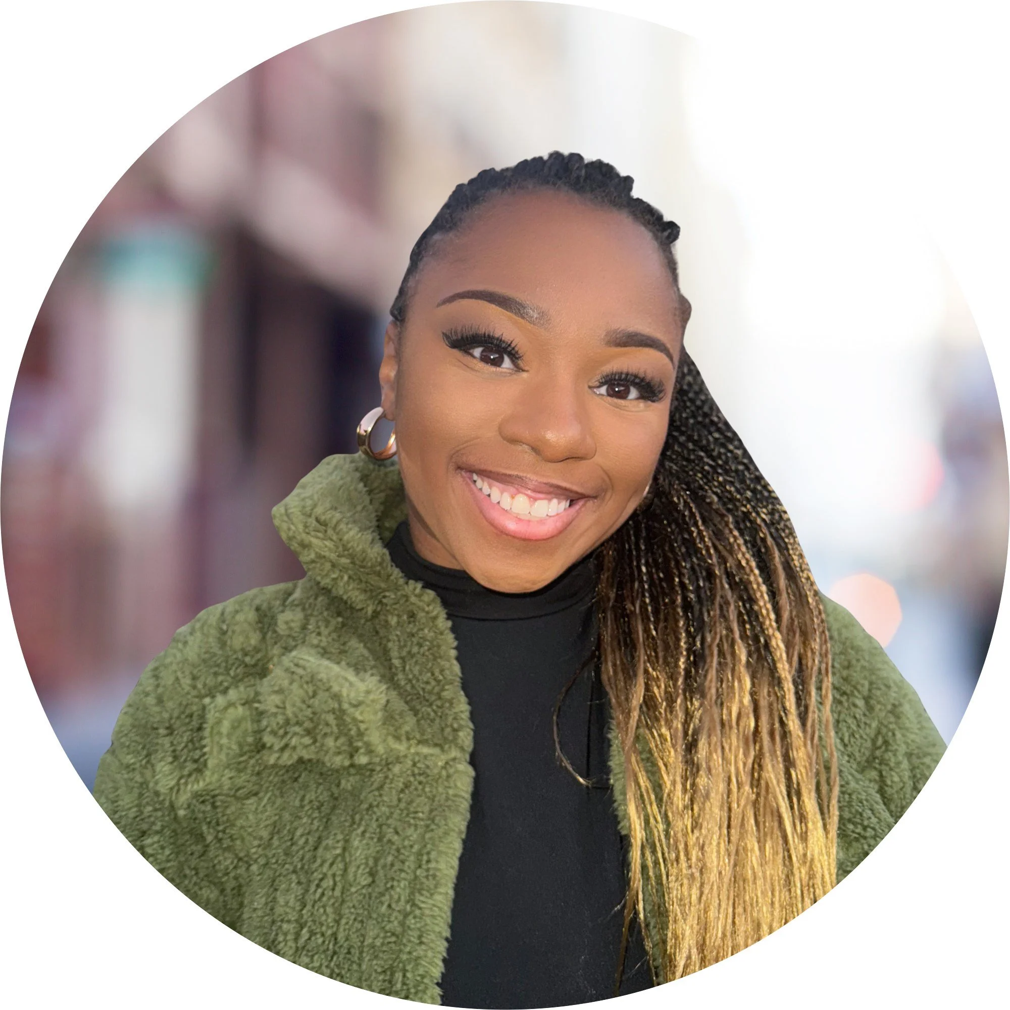 Close-up of a smiling woman with braided hair, wearing gold hoop earrings and a green textured jacket.