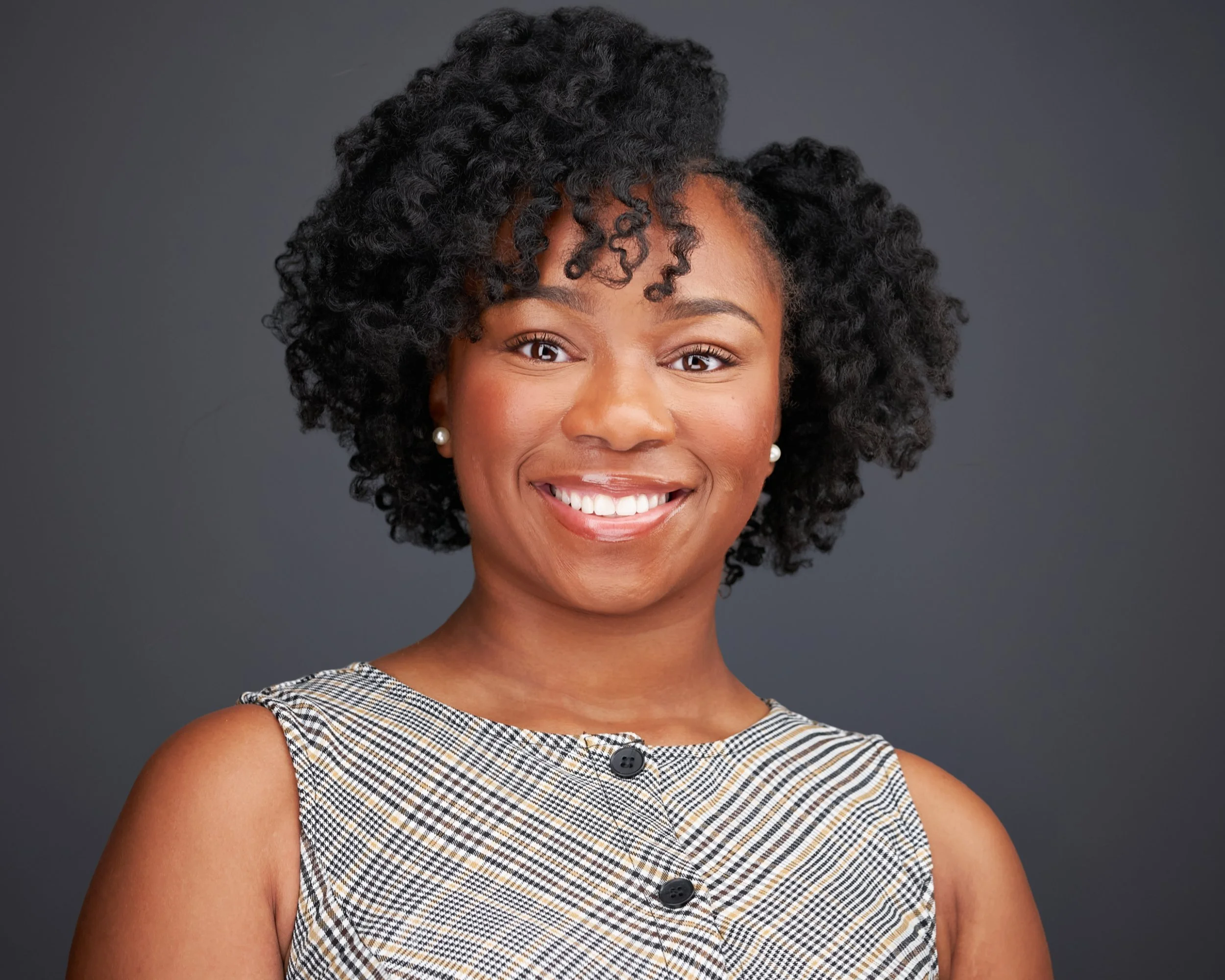 Close-up of an African American woman smiling, with short, curly black hair and pearly earrings, wearing a sleeveless plaid top against a plain gray background.