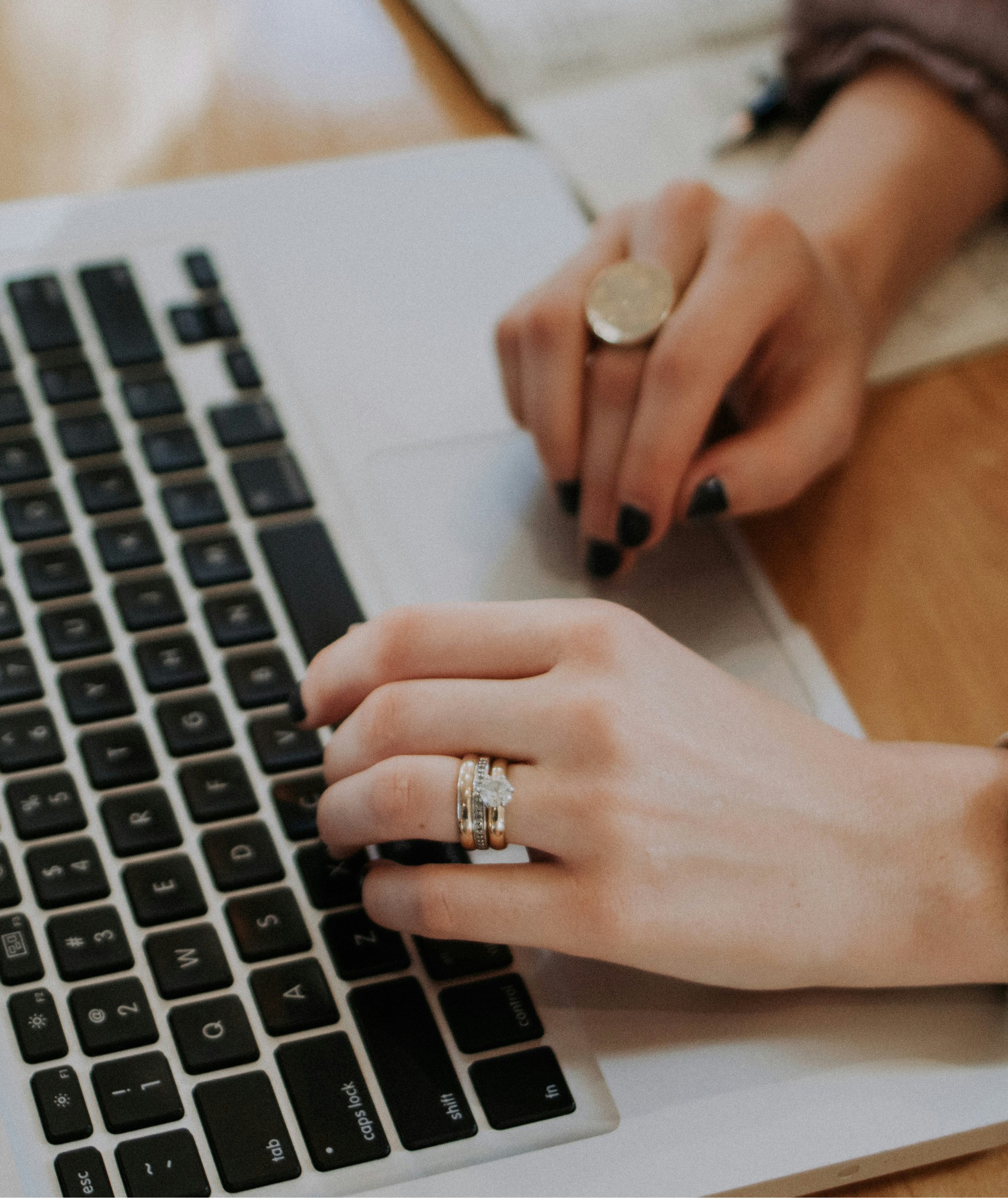 Close-up of a person’s hand resting on a laptop keyboard, wearing multiple rings, including a large ring on the middle finger and an engagement ring with a diamond on the ring finger.