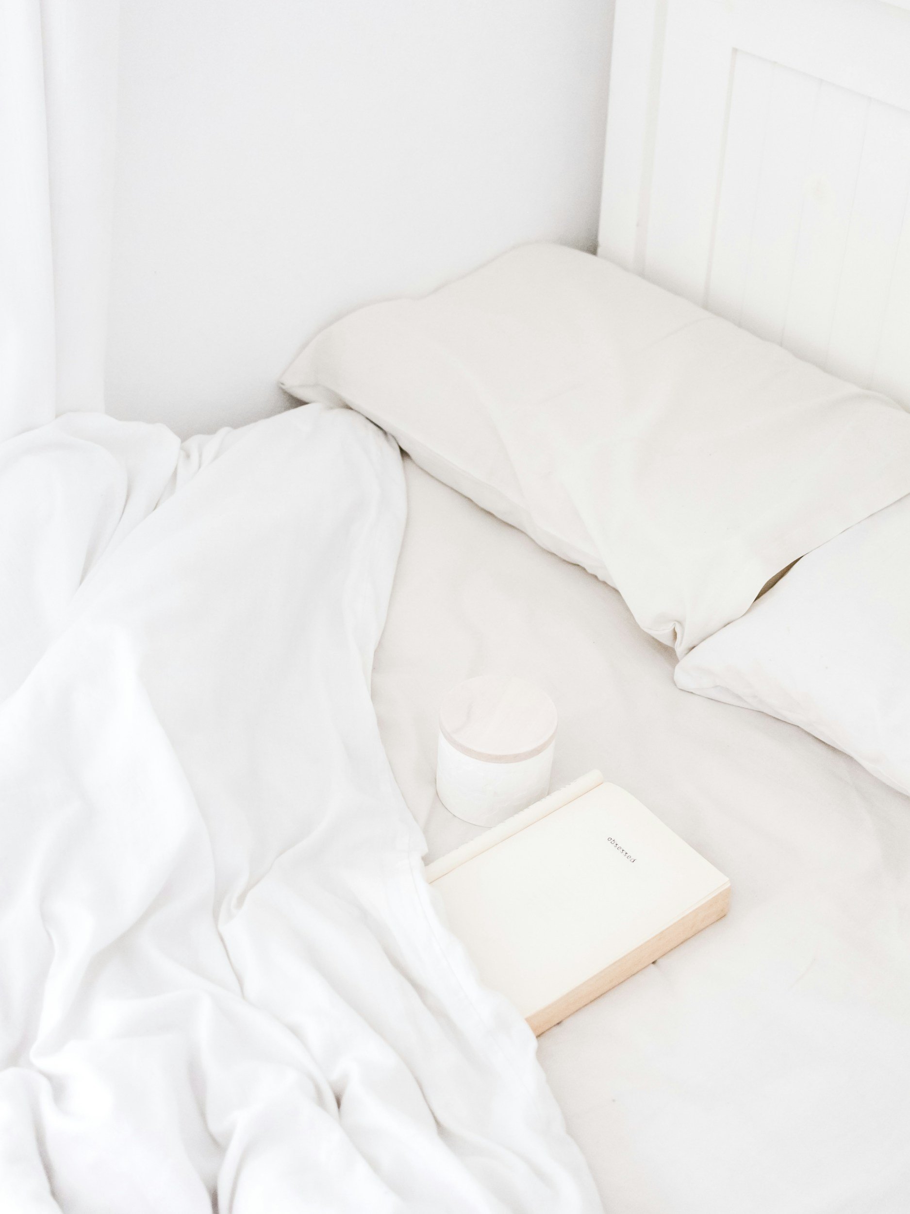 A minimalistic bed setup with white linens, two pillows, a book titled 'Passed', a can of beverage, and a white ceramic cup placed on a bed with white sheets and a white headboard.