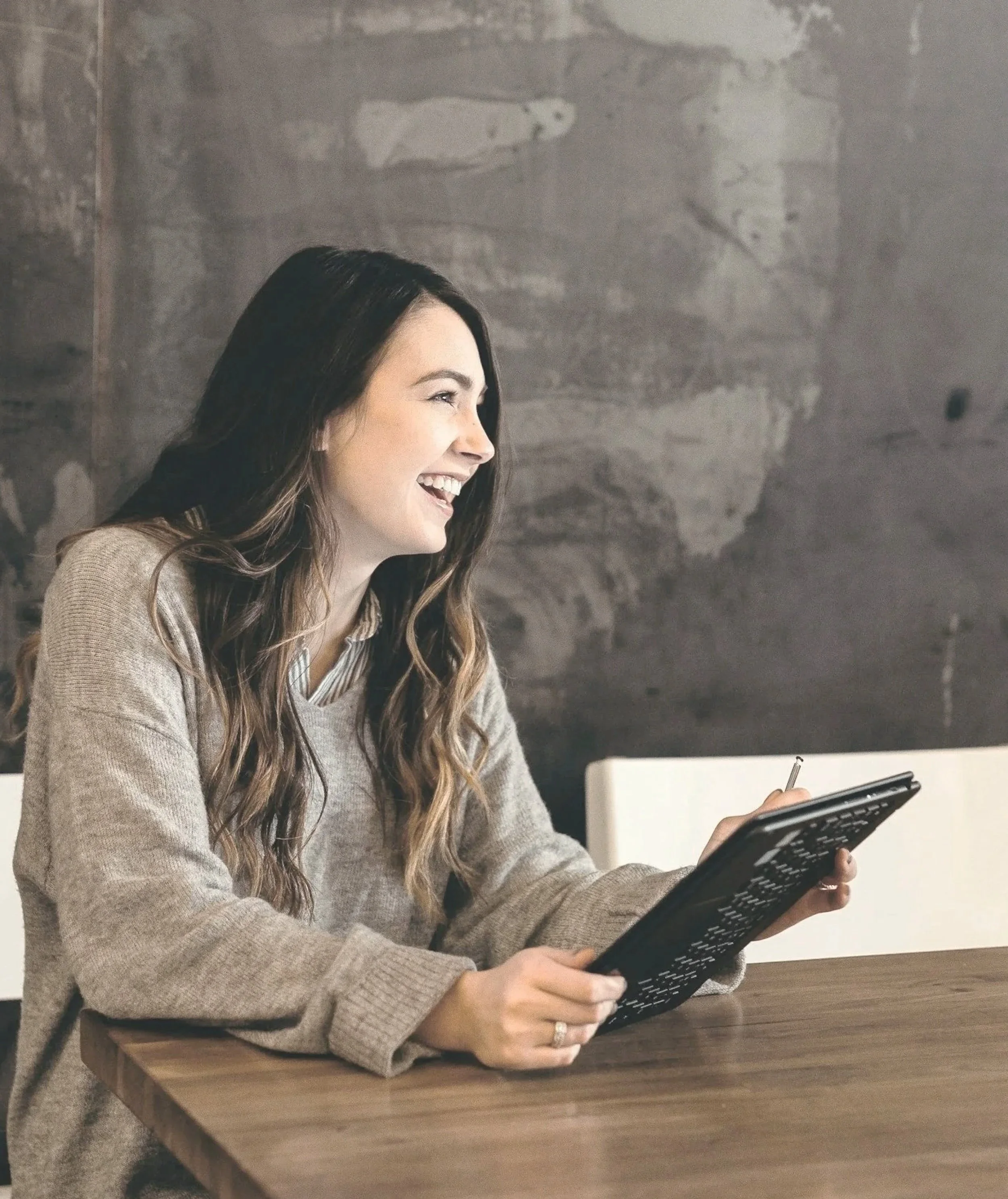 A woman with long dark hair smiling and looking to her right while holding a tablet device in her right hand, seated at a wooden table with a gray textured wall behind her.