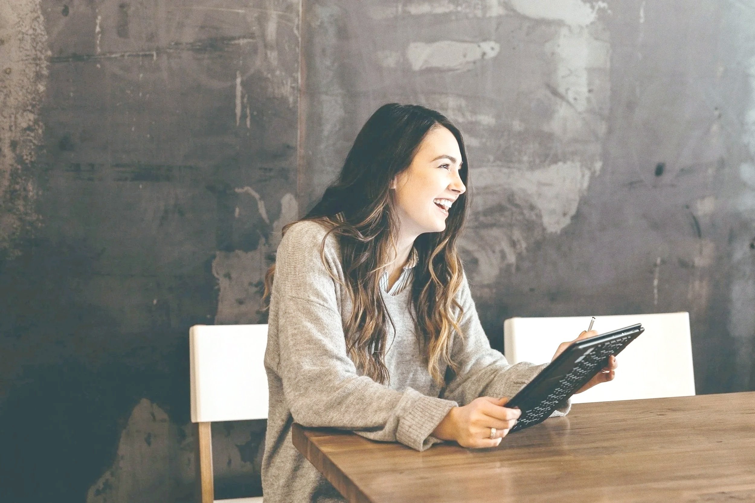 A woman with long dark hair smiling and looking to her right while sitting at a wooden table, holding a tablet device in her left hand, in a room with a gray textured wall.