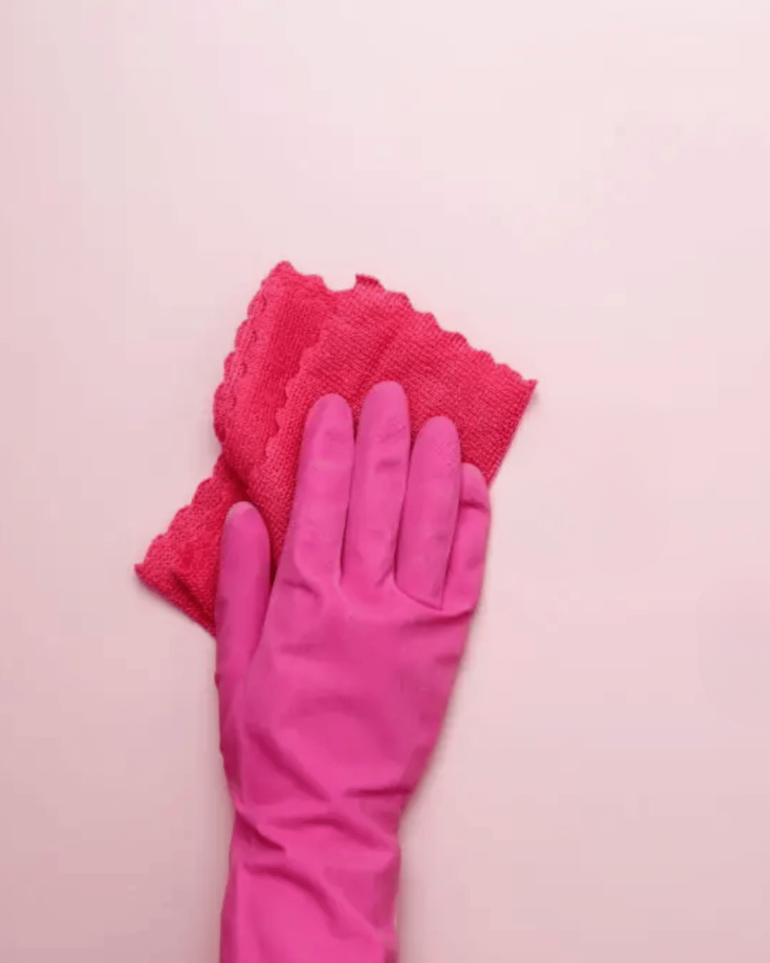 A hand wearing a pink rubber glove holding a folded pink cleaning cloth on a white background.