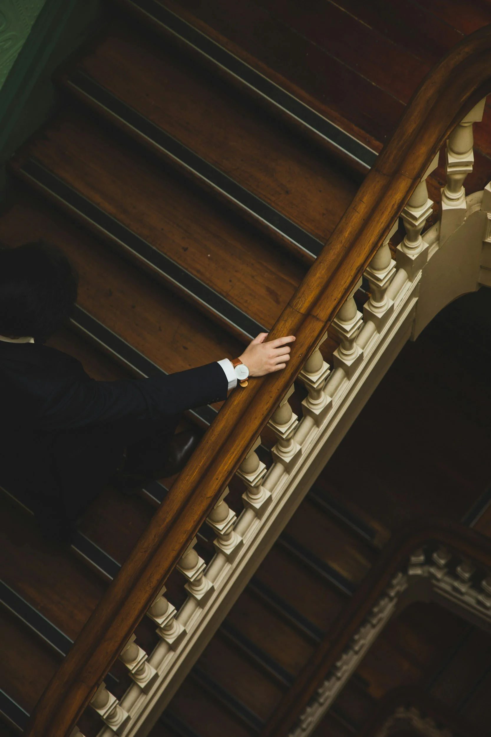 Sem Gulinyan with a watch on his wrist is holding a wooden handrail while walking down a staircase with dark wooden steps and white balusters.