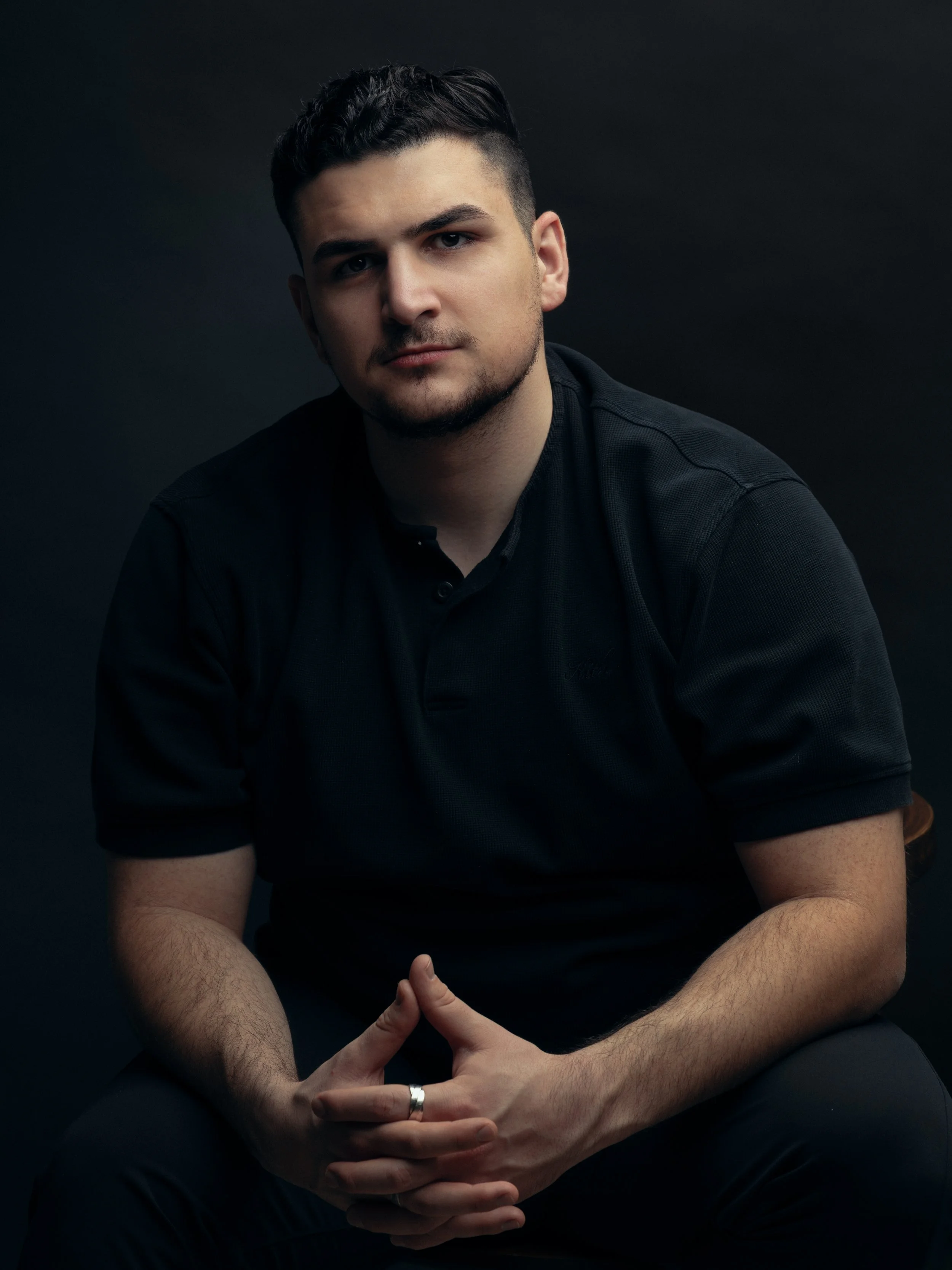 Portrait of a young man; Sem Gulinyan with dark hair, sitting against a dark background, wearing a black shirt, with hands clasped and a small smile.