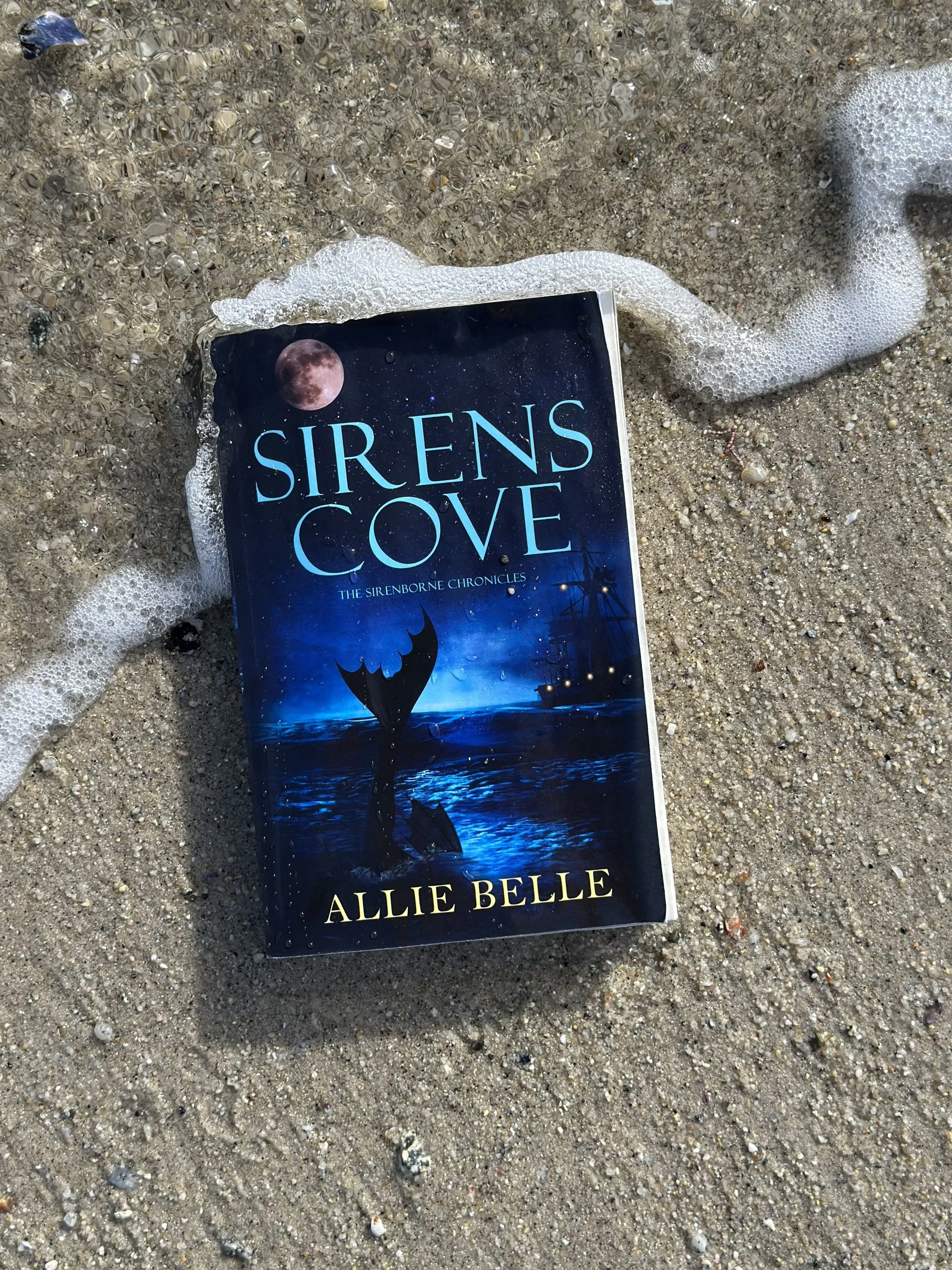 A book titled 'Sirens Cove: The Sirenborne Chronicles' by Allie Belle lies face down on a sandy beach, partially submerged in the sand with foamy surf surrounding it.
