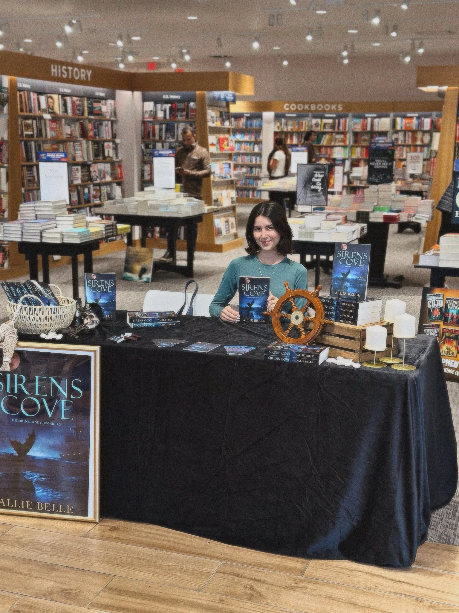 A woman sits at a book signing table in a bookstore, promoting her book titled 'Sirens Cove' by Allie Belle. The table displays several copies of the book, a nautical-themed ship wheel, candles, and promotional materials, with shelves of books in the background.