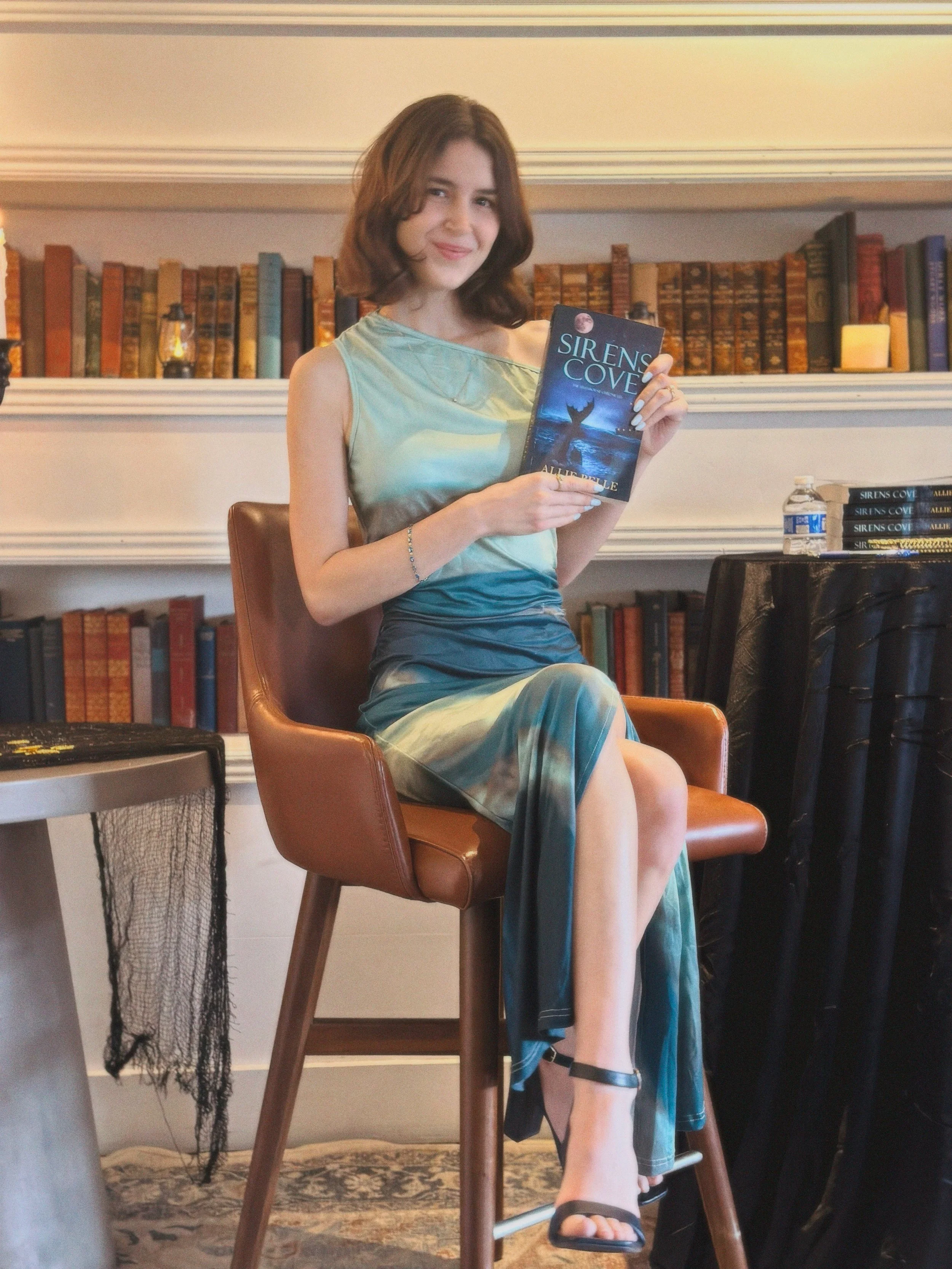 A woman sitting on a wooden chair, holding a book titled 'Sirens Cove' by Allie Belle, in a cozy room with a bookshelf, a water bottle, and a candle in the background.