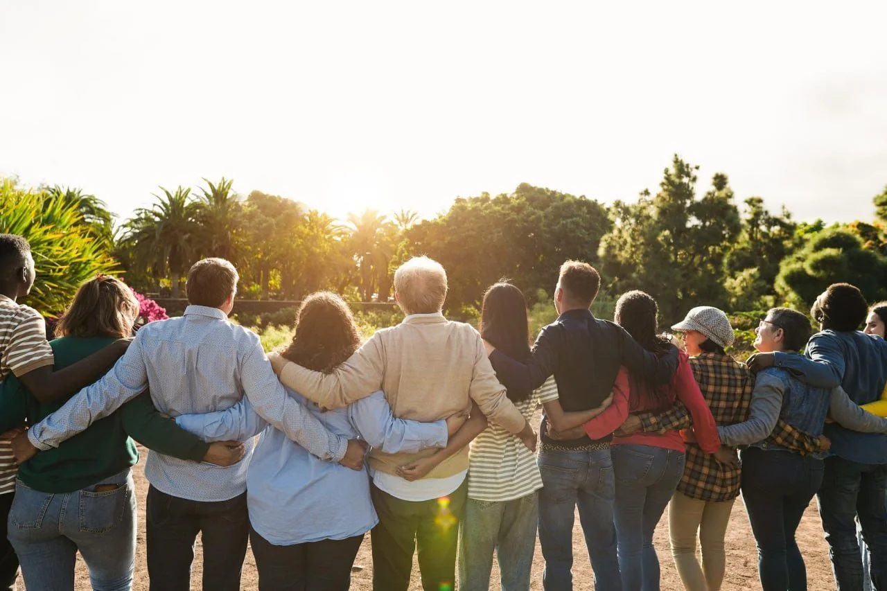 Group of diverse people standing outdoors with their arms around each other during sunset, facing away from the camera.
