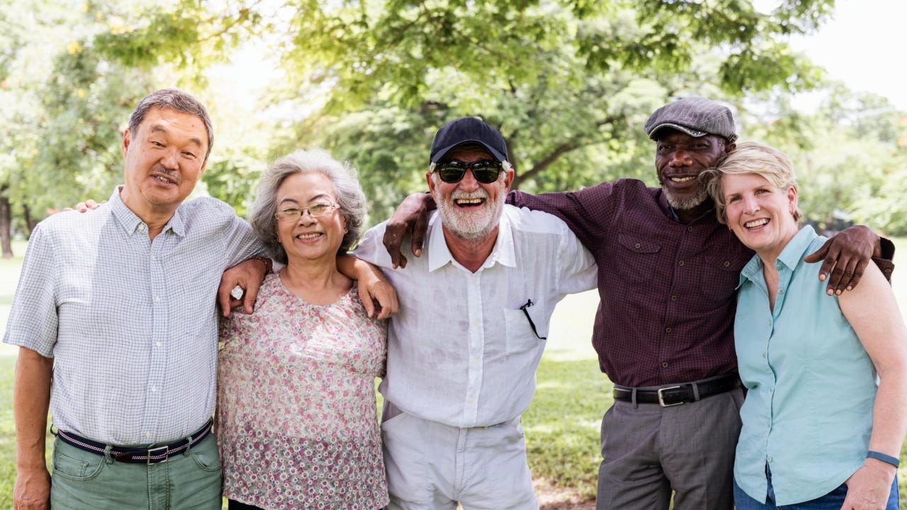 Group of five diverse people standing outdoors with trees in the background, smiling and embracing each other.