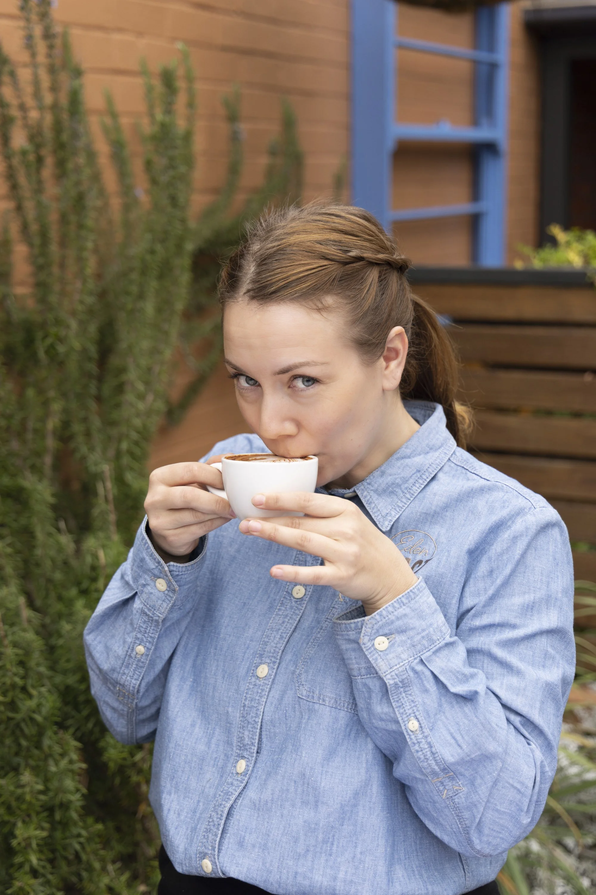 A woman with light brown hair in a braid, wearing a denim shirt, drinks coffee outdoors. There is a wooden fence and green plants in the background.