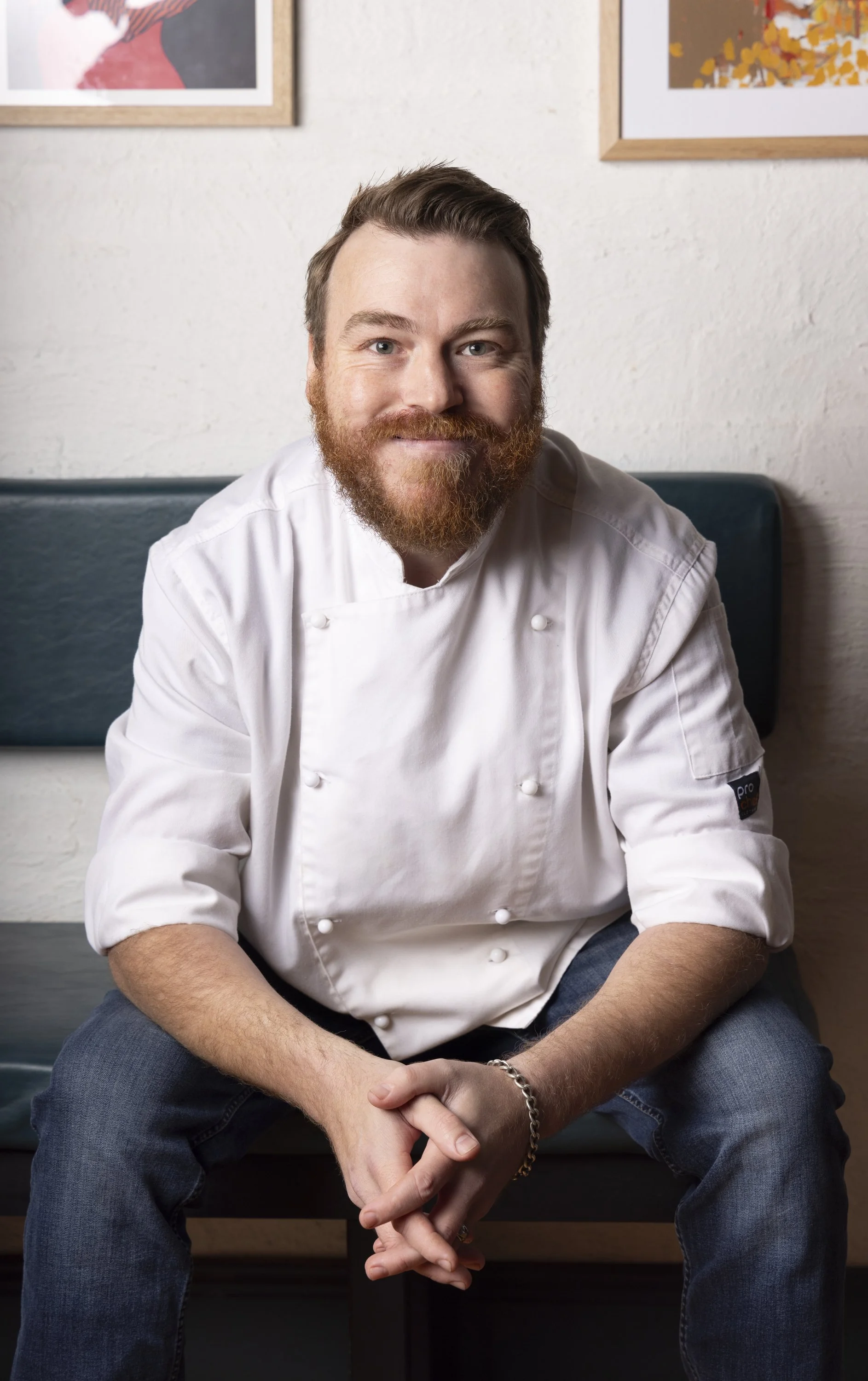 A smiling male chef seated on a bench, wearing a white chef coat with rolled-up sleeves and blue jeans, in a room with framed artwork on a white wall.