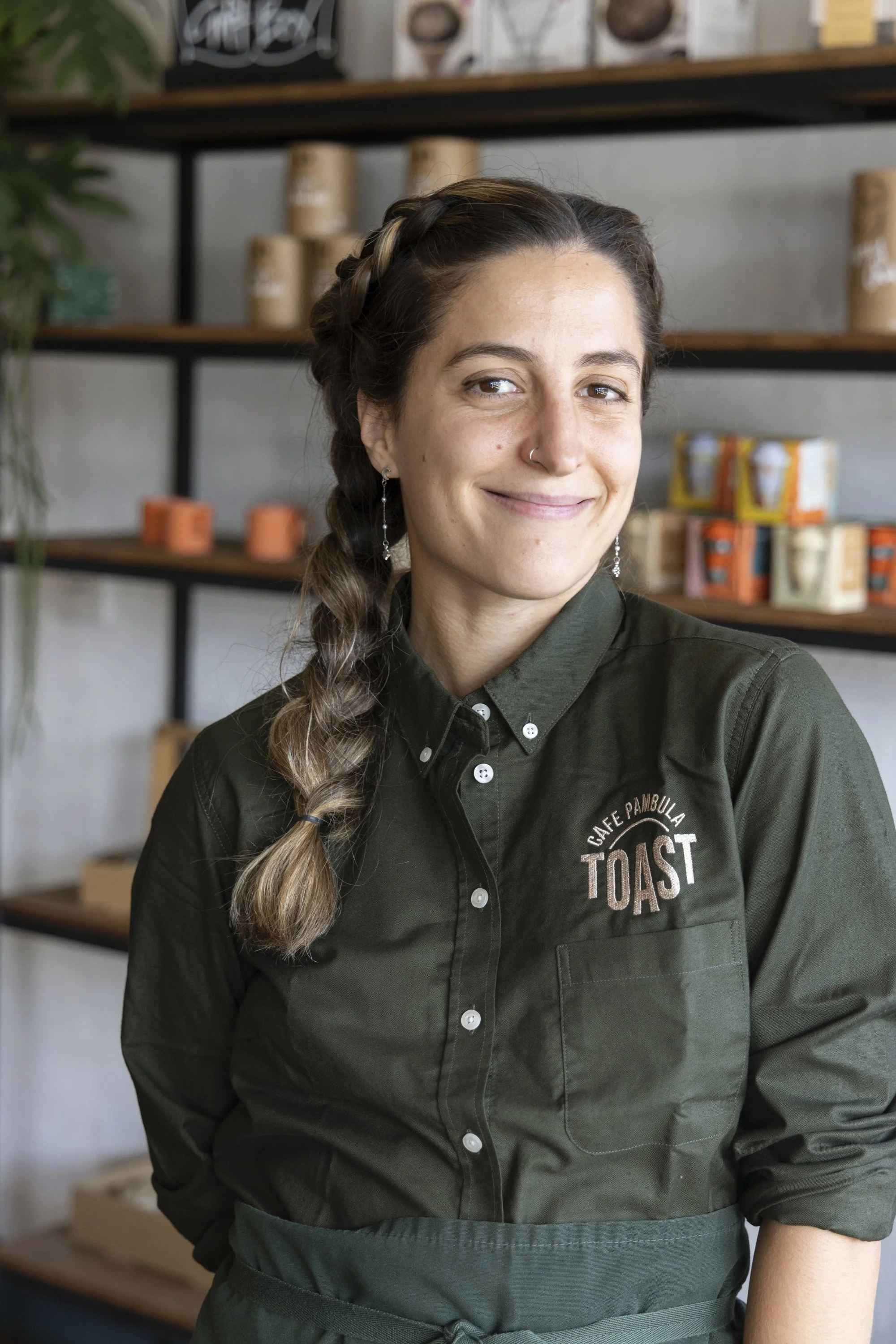 A woman wearing a dark green collared shirt with a cafe logo, standing inside a coffee shop with shelves of coffee products behind her.
