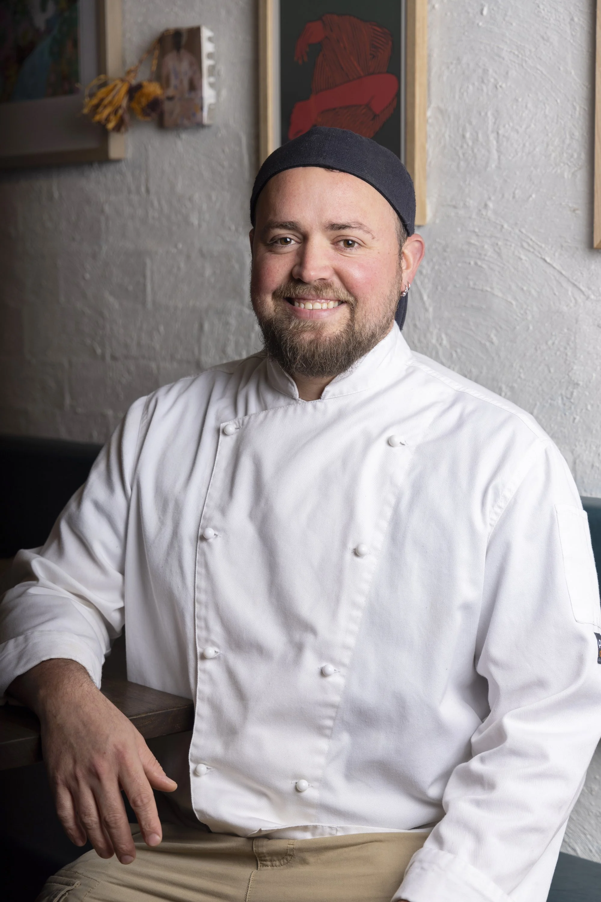 A smiling male chef with a beard and earring, wearing a white chef's coat and a black hat, sitting in a restaurant with artwork on the wall behind him.