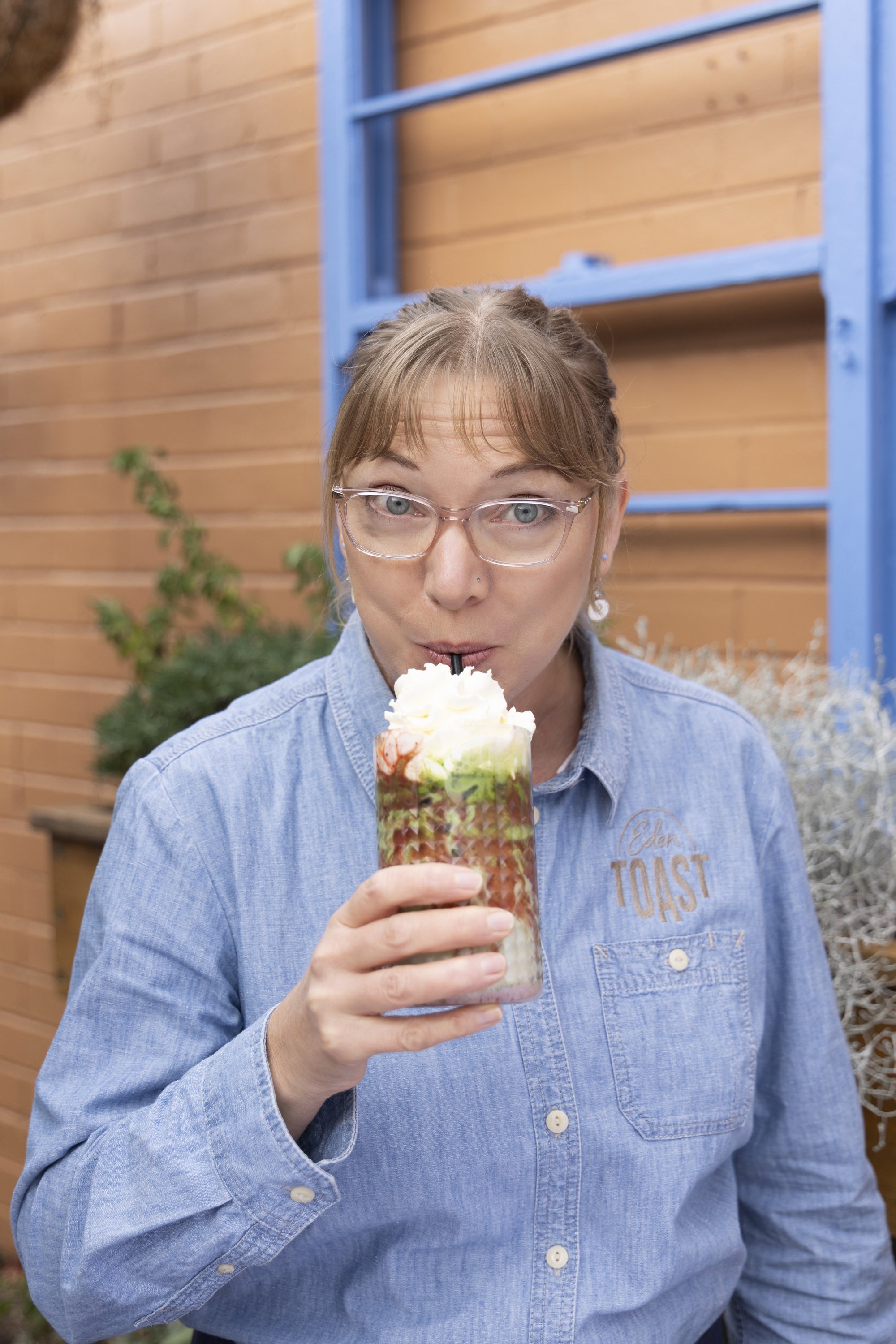 A woman wearing glasses and a denim shirt with 'Eden Toast' embroidered on it is enjoying a tropical drink topped with whipped cream outdoors, against a background of a wooden wall, a blue ladder, and some plants.