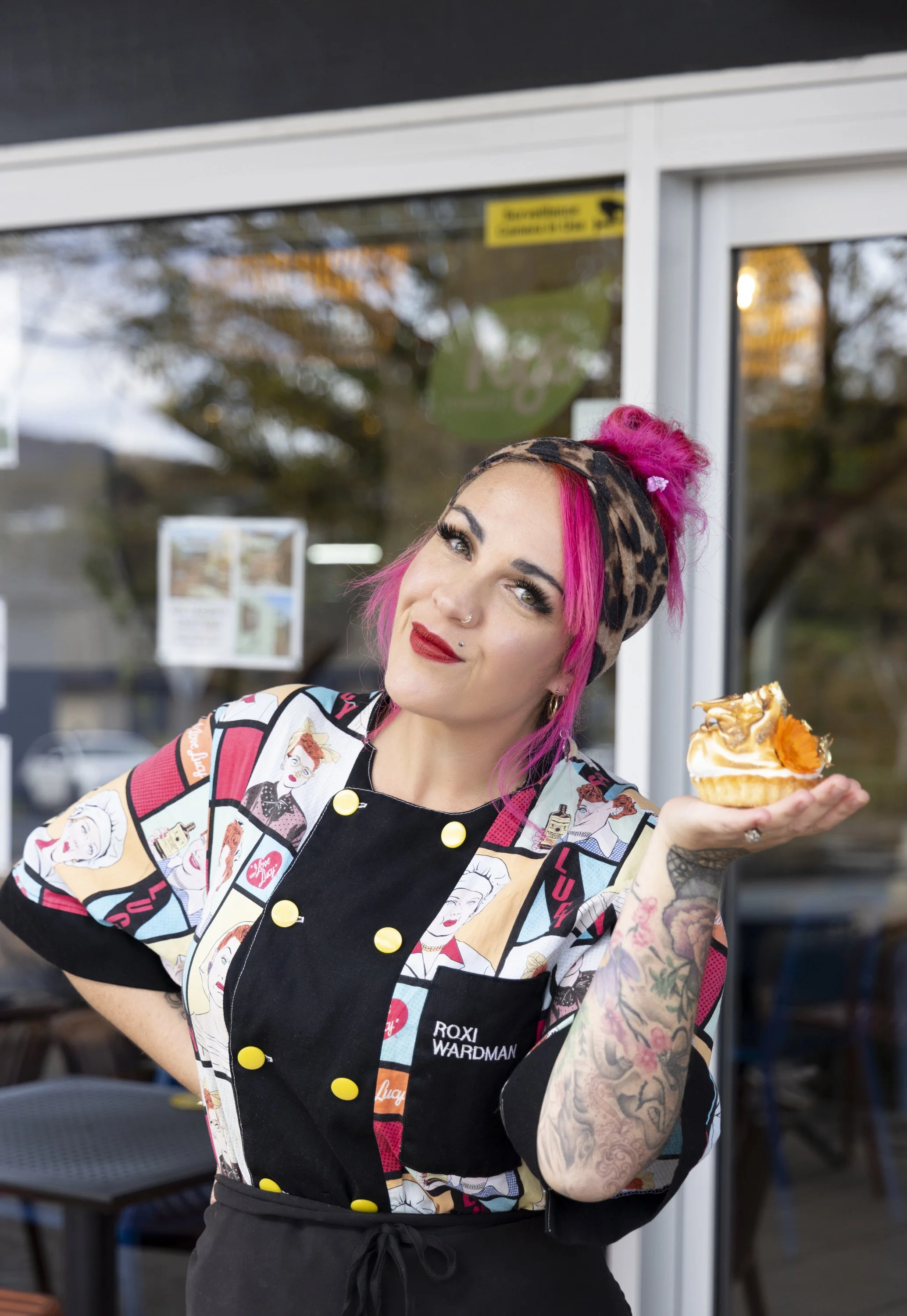 A woman with bright pink hair, tattoos, and makeup, wearing a patterned chef's coat labeled 'Roxi Wardman,' holding a cupcake with caramel and orange decorations outside a café.