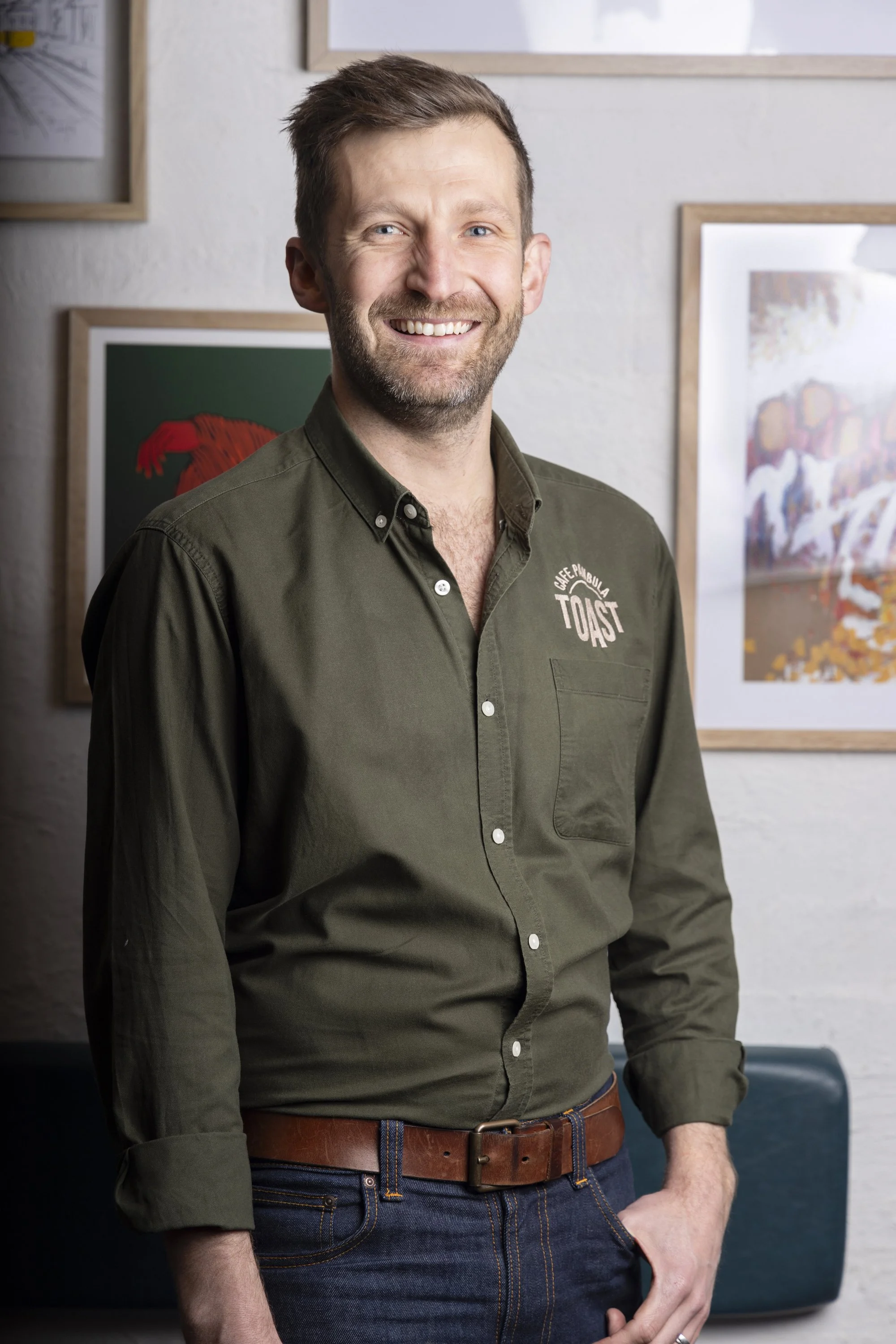 A smiling man with brown hair and a beard, wearing a green button-up shirt with 'Toast' written on it, standing in front of a wall with framed artwork.