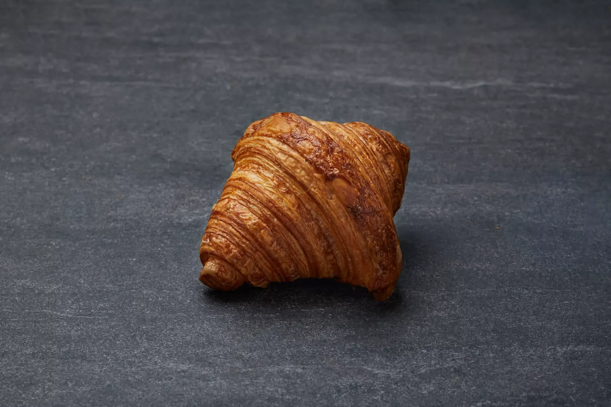 A single, golden brown, flaky croissant on a dark, textured surface.
