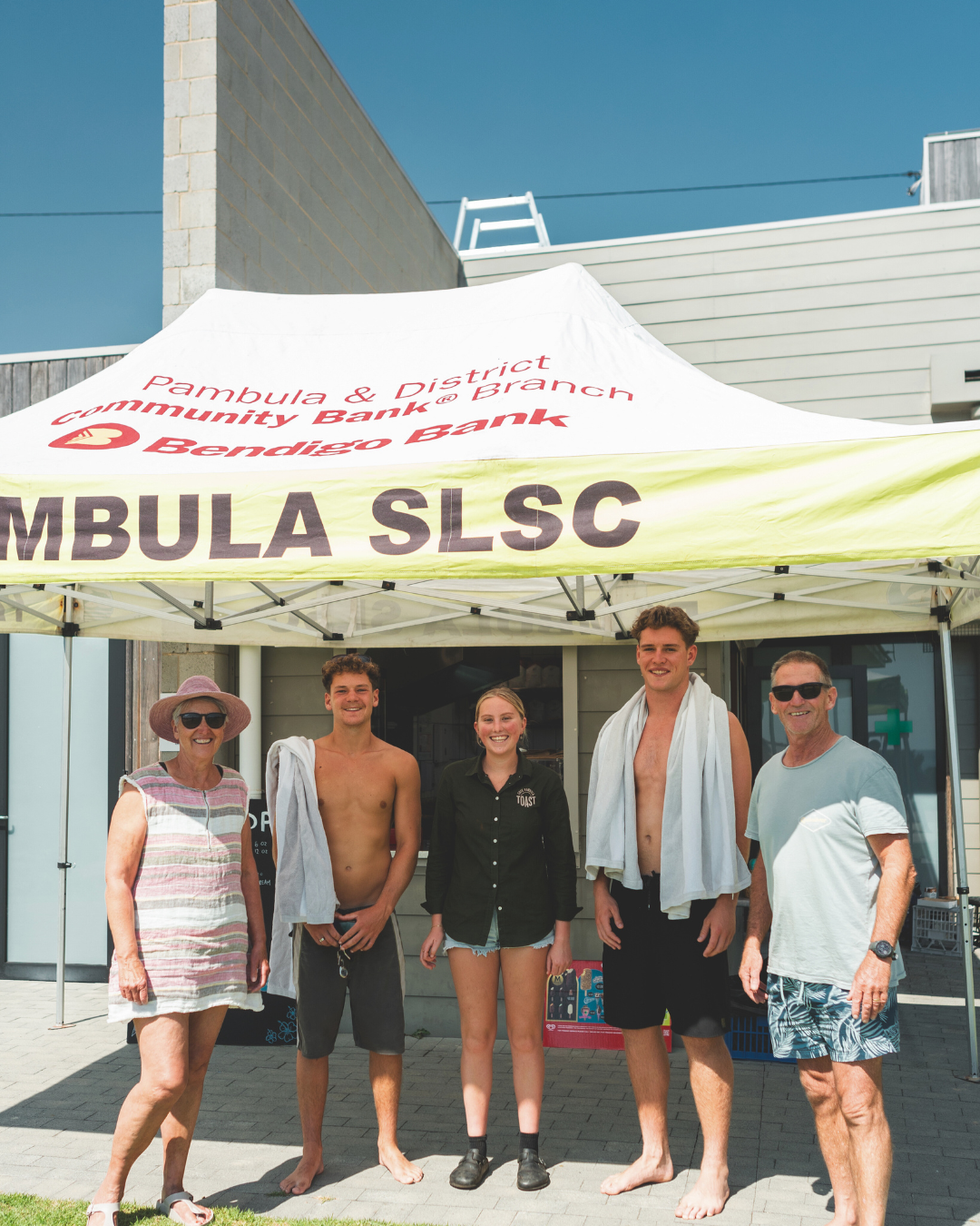 Group of five people standing under a white canopy tent with red and yellow text, outdoors on a sunny day