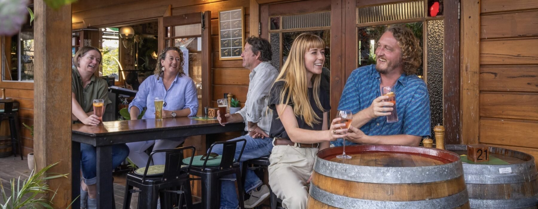 Group of five friends laughing and enjoying drinks at an outdoor bar with wooden walls, two barrels, and large windows.