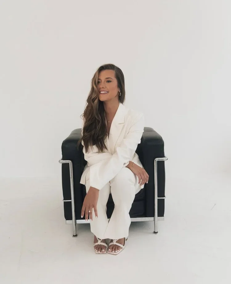 A woman with long wavy hair sitting on a black chair against a plain white background, smiling and wearing a white suit and white strappy heels.