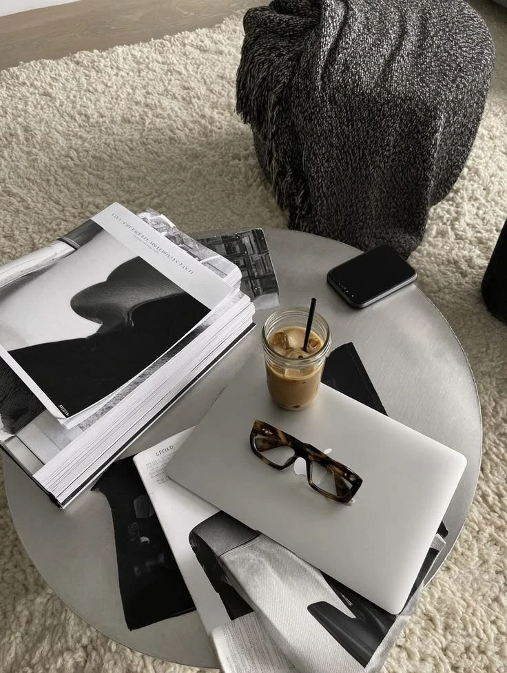 A round coffee table with magazines, a glass of iced coffee, a pen, a closed laptop with sunglasses on top, a smartphone, and a black booklet. A gray furry chair and a textured black blanket are visible near a cream-colored rug.