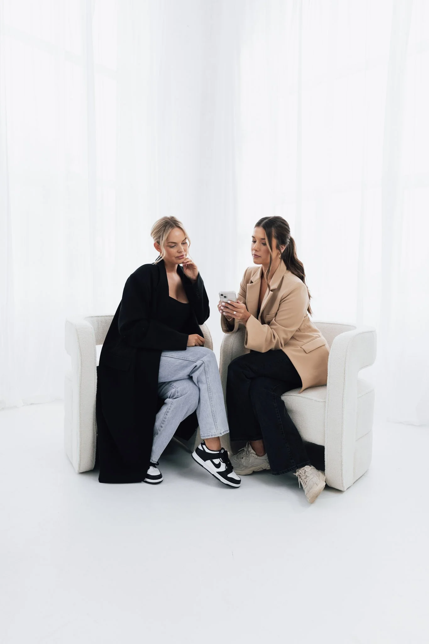 Two women sitting on white chairs against a white background, one showing her phone to the other.