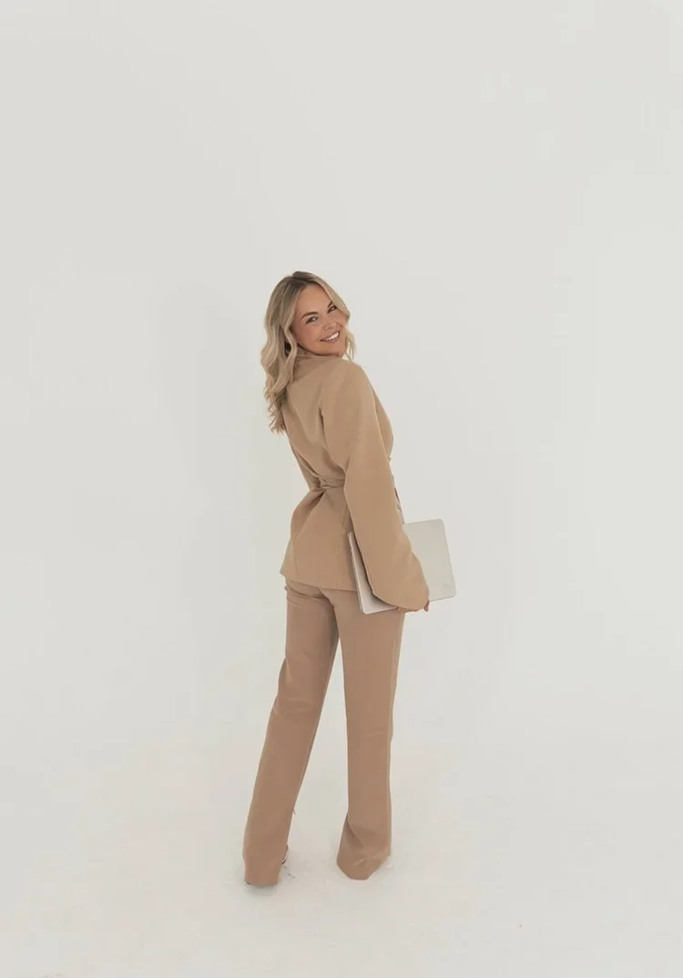 A woman in a beige suit holding a laptop, smiling, against a white background.
