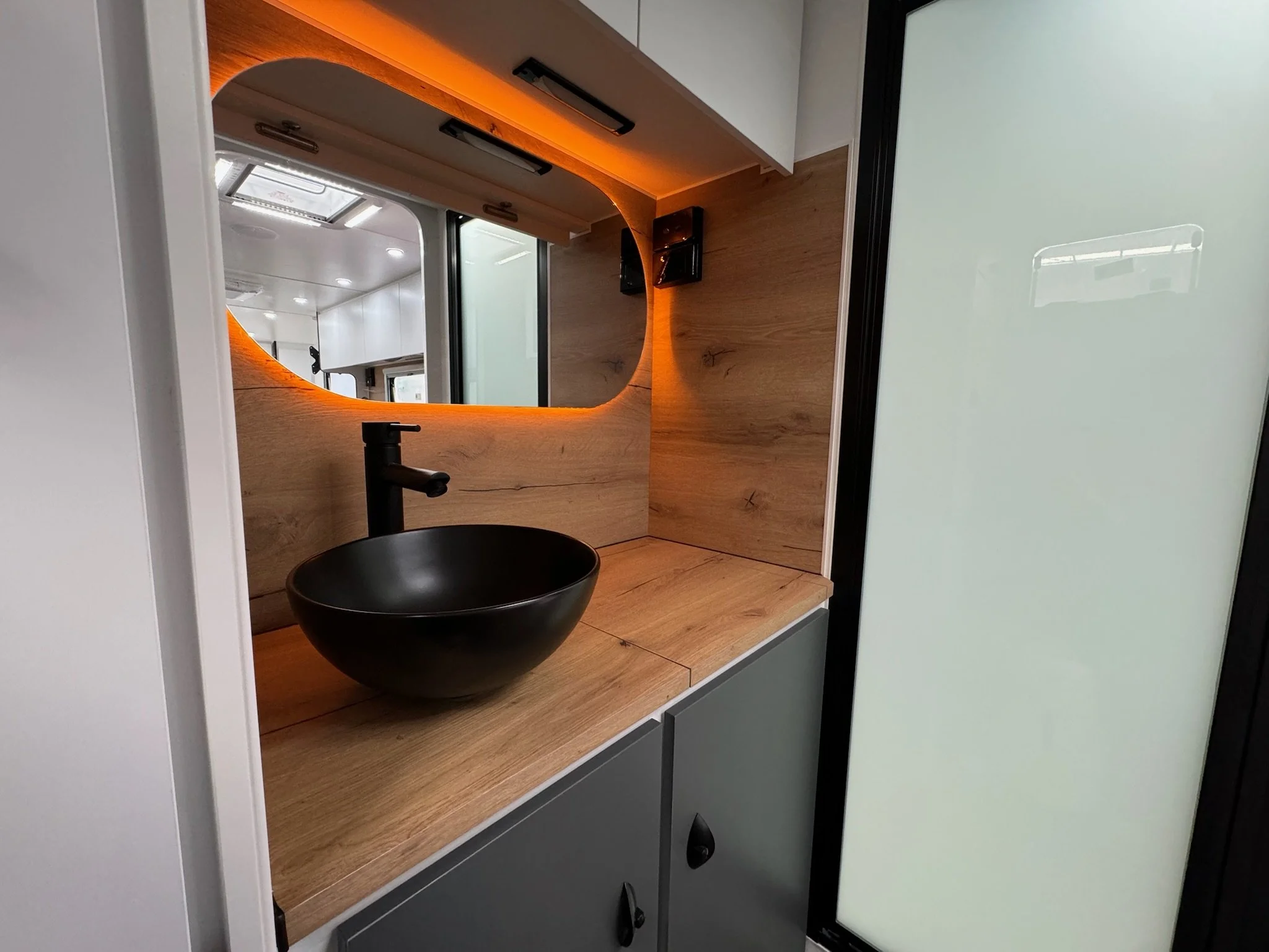 A modern bathroom vanity with a black vessel sink, black faucet, and warm wood accents. A large mirror with orange backlighting is mounted above the sink, and there are white cabinets and a frosted glass panel visible.