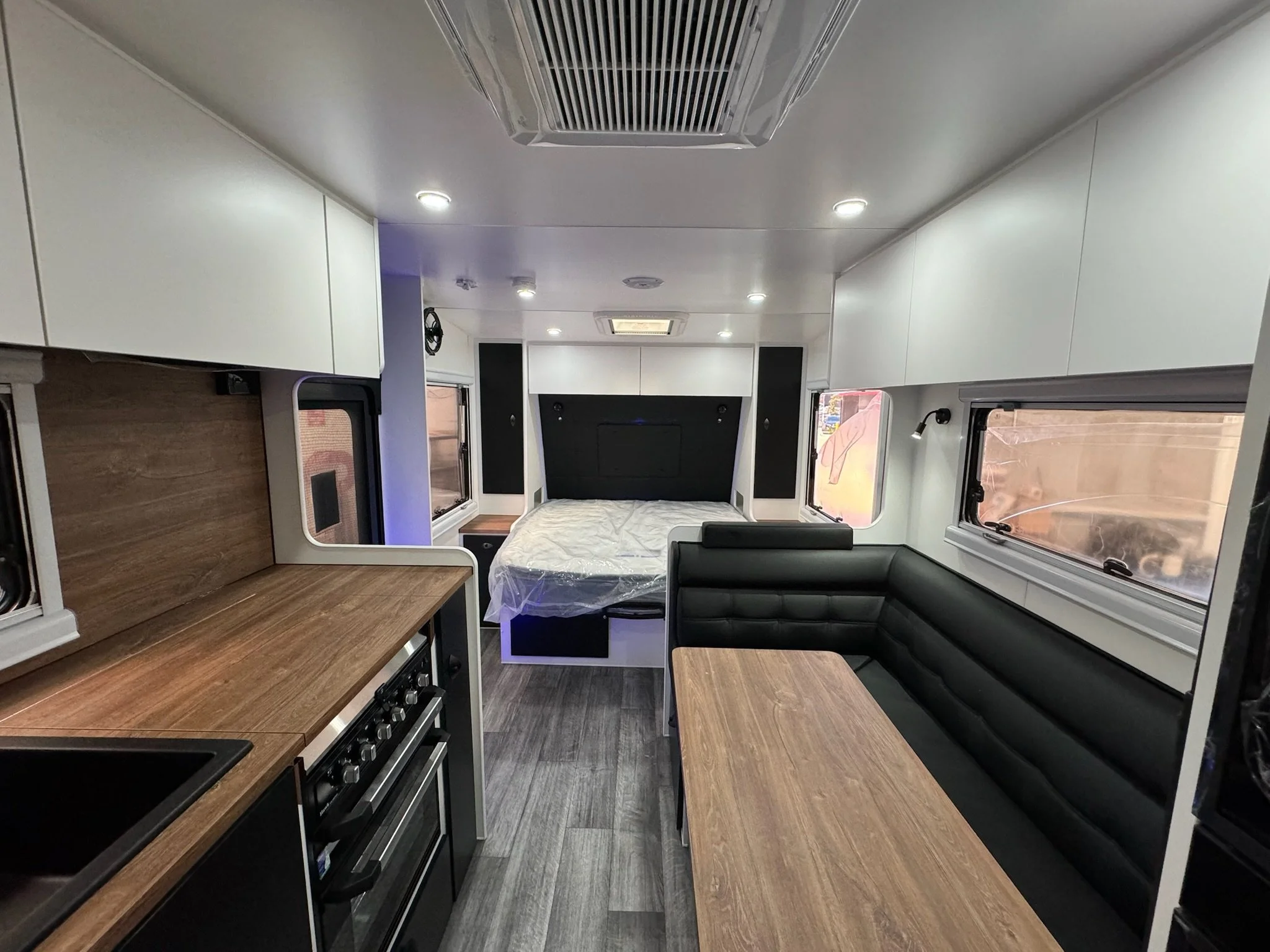 Interior of a modern camper van showing a kitchen area with wooden countertops, a stove, and white cabinets, with a bed and windows visible at the back.