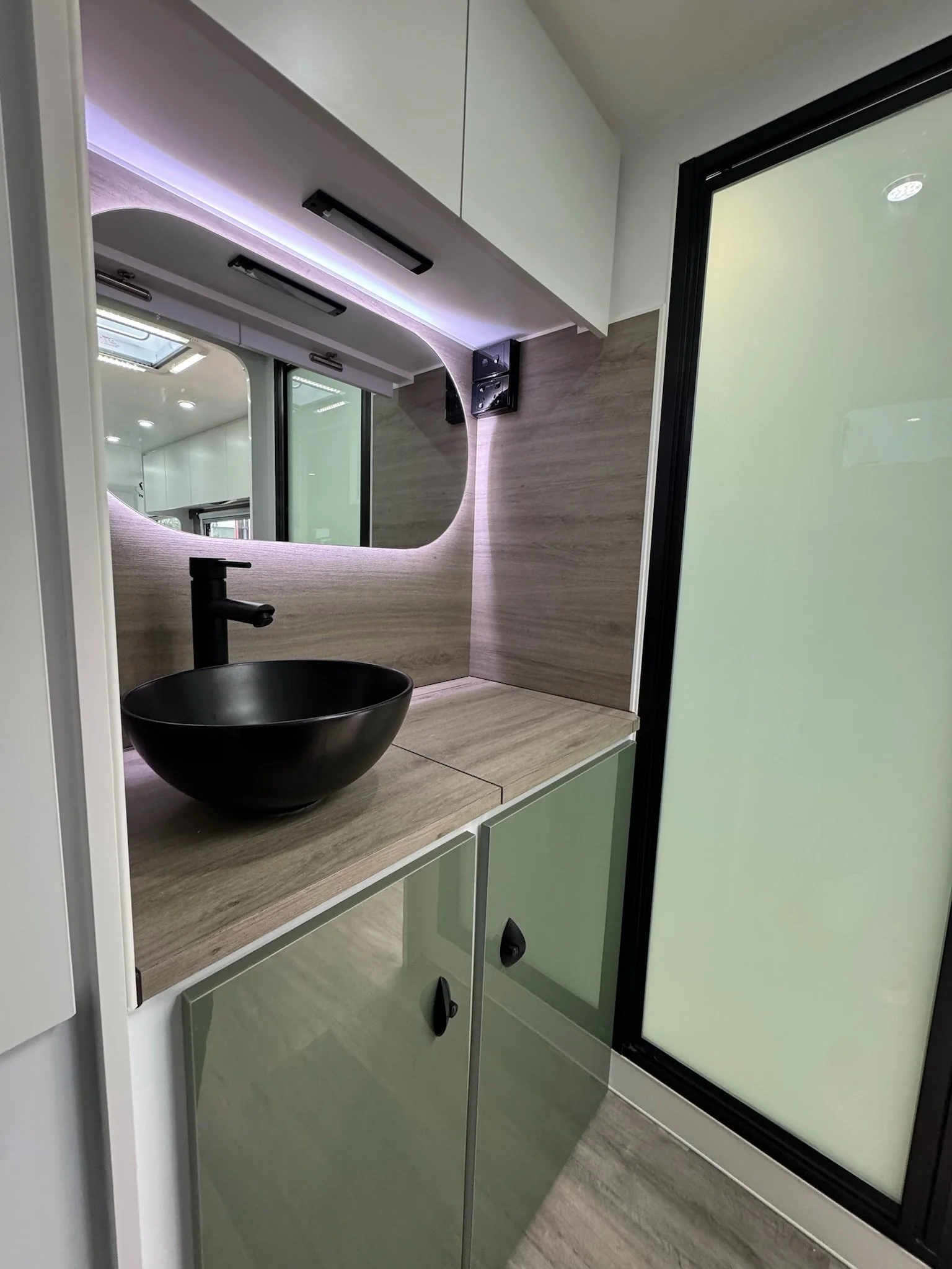 Modern bathroom with a black round vessel sink, wooden countertop, light green cabinets, a mirror, and sliding frosted glass door.