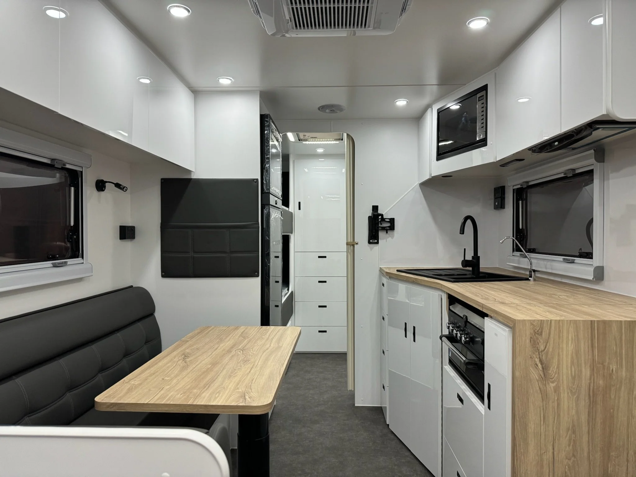 Interior of a tiny home or camper with white cabinetry, a small microwave, black faucet, and a wooden countertop adjacent to a seating area with a black upholstered bench and a wooden table. There are small windows and ceiling lights.