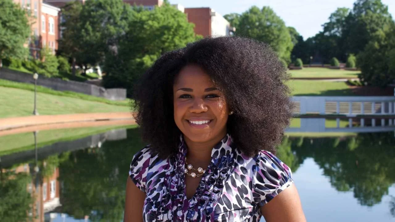A woman with natural, curly hair standing outdoors near a pond with trees and buildings in the background.