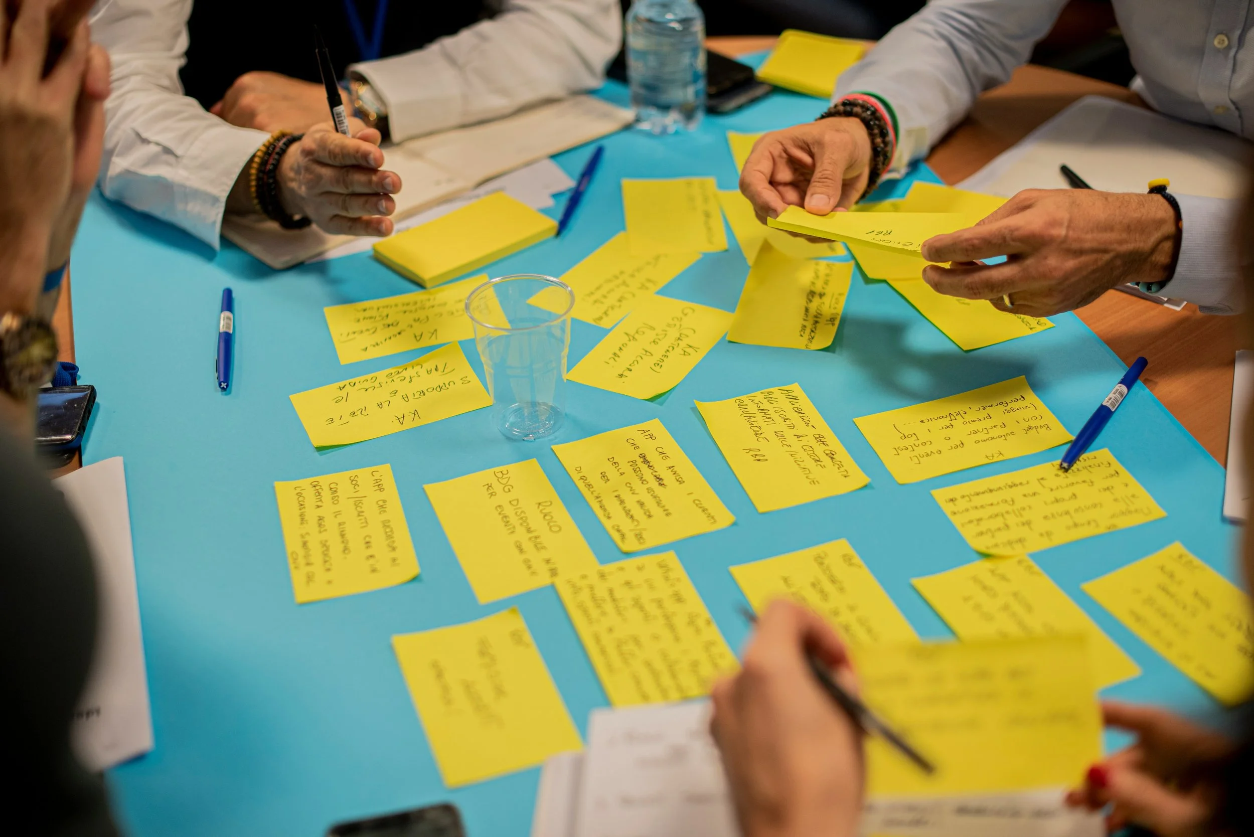 Multiple people are working together around a table with bright yellow notes, pens, and notebooks, collaborating and sharing ideas.