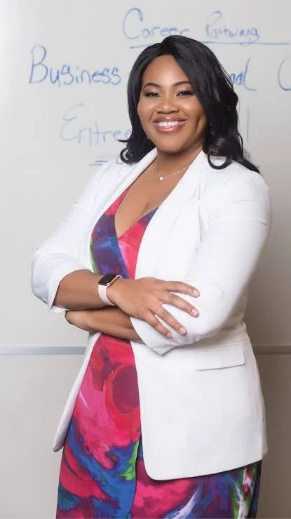 A smiling woman with dark hair, wearing a white blazer over a colorful dress, standing with arms crossed in front of a whiteboard with notes related to business.