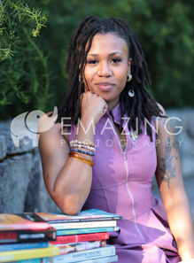 A woman with long dreadlocks sitting outdoors at a table with stacks of books, wearing a purple dress and bracelets, with trees in the background.