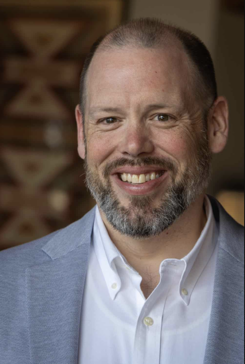 A smiling man with a beard, wearing a white shirt and light blue blazer, in an indoor setting with wooden background.