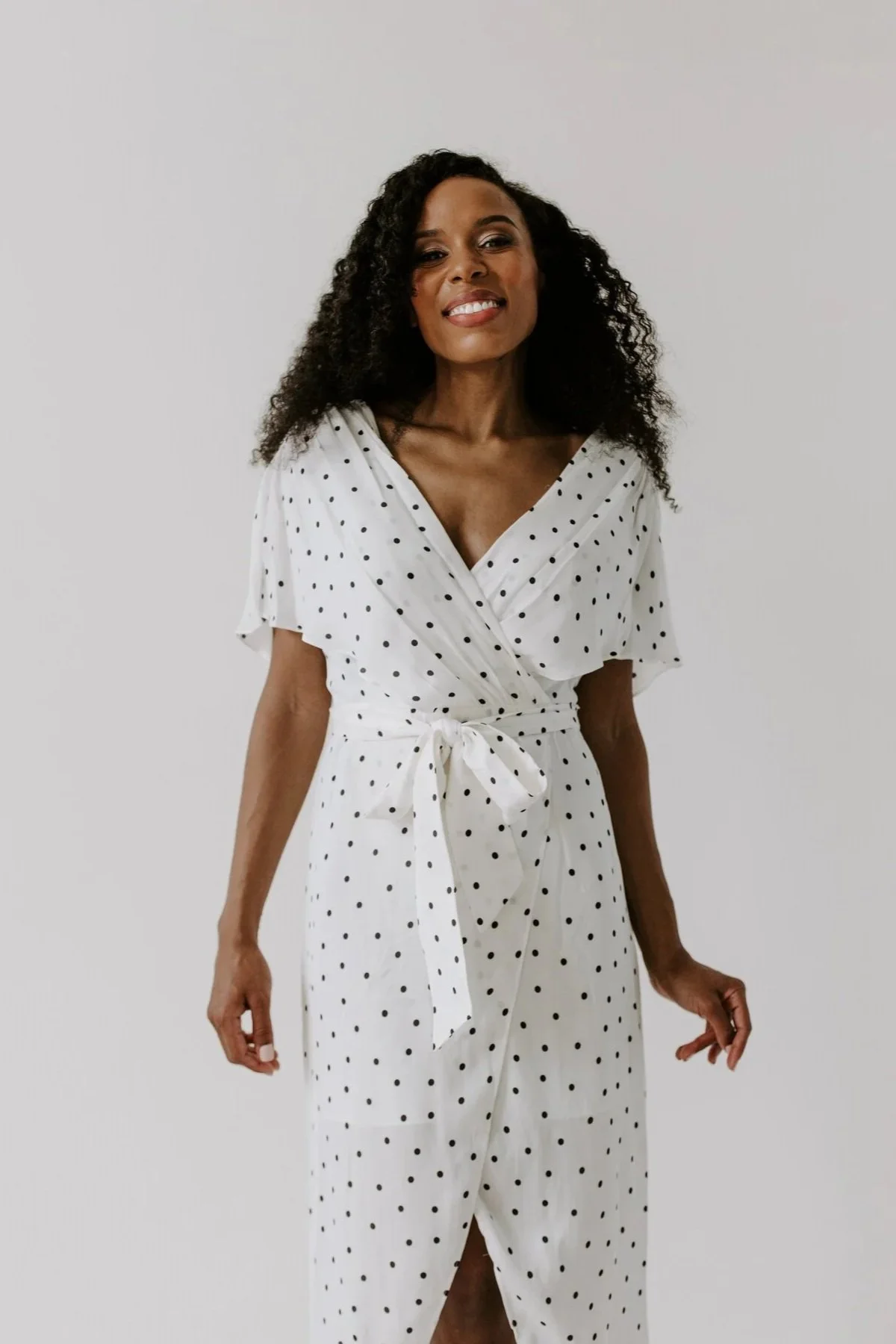A woman wearing a white dress with black polka dots, tied at the waist, standing against a plain background and smiling.